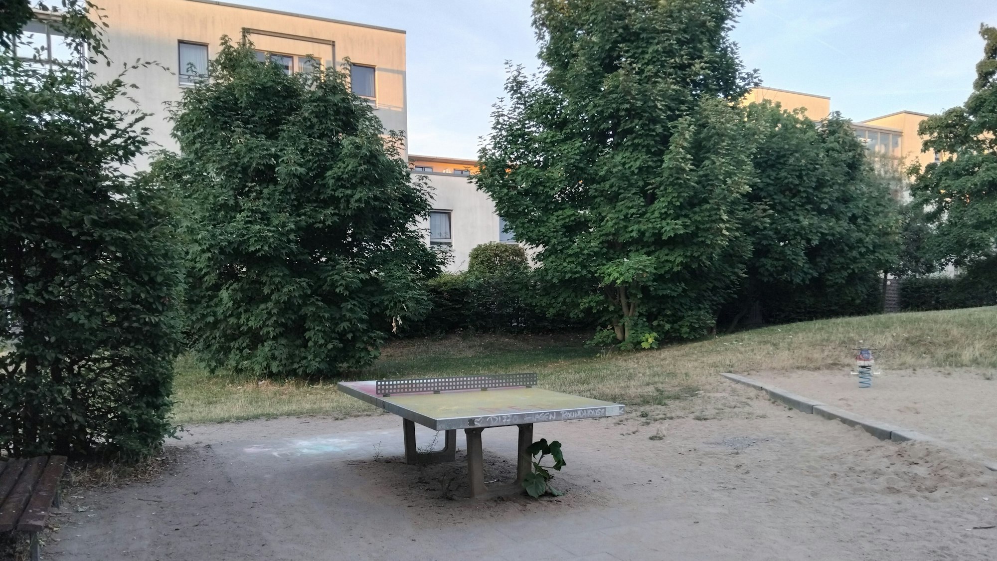 Tischtennisplatte neben einem Spielplatz, im Hintergrund Bäume und Wohngebäude.