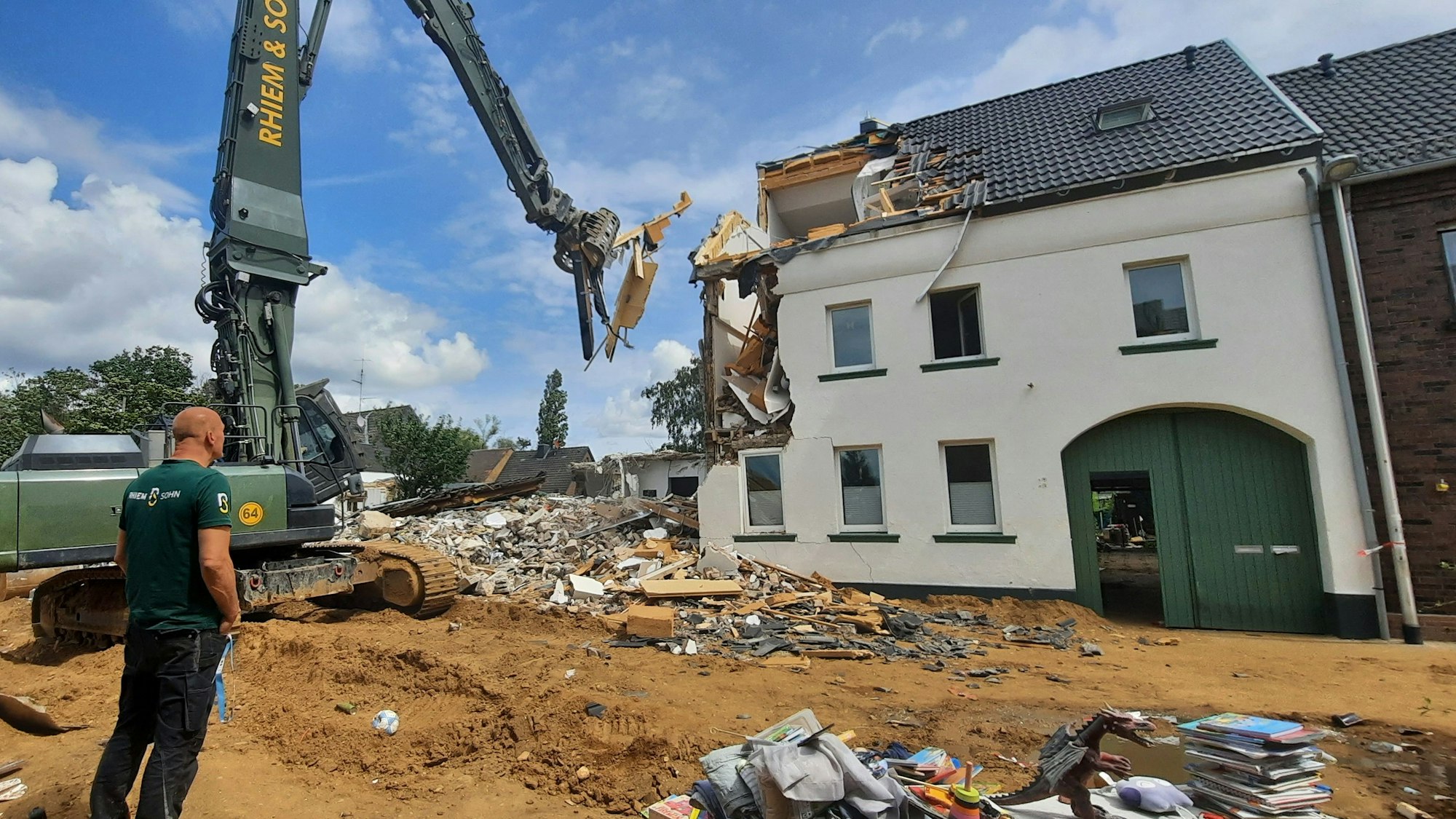 Zu sehen ist ein Haus, das von einem Bagger abgerissen wird, davor steht ein Mann.