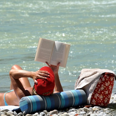 Eine Frau liest am Ufer ein Buch im Sonnenschein.
