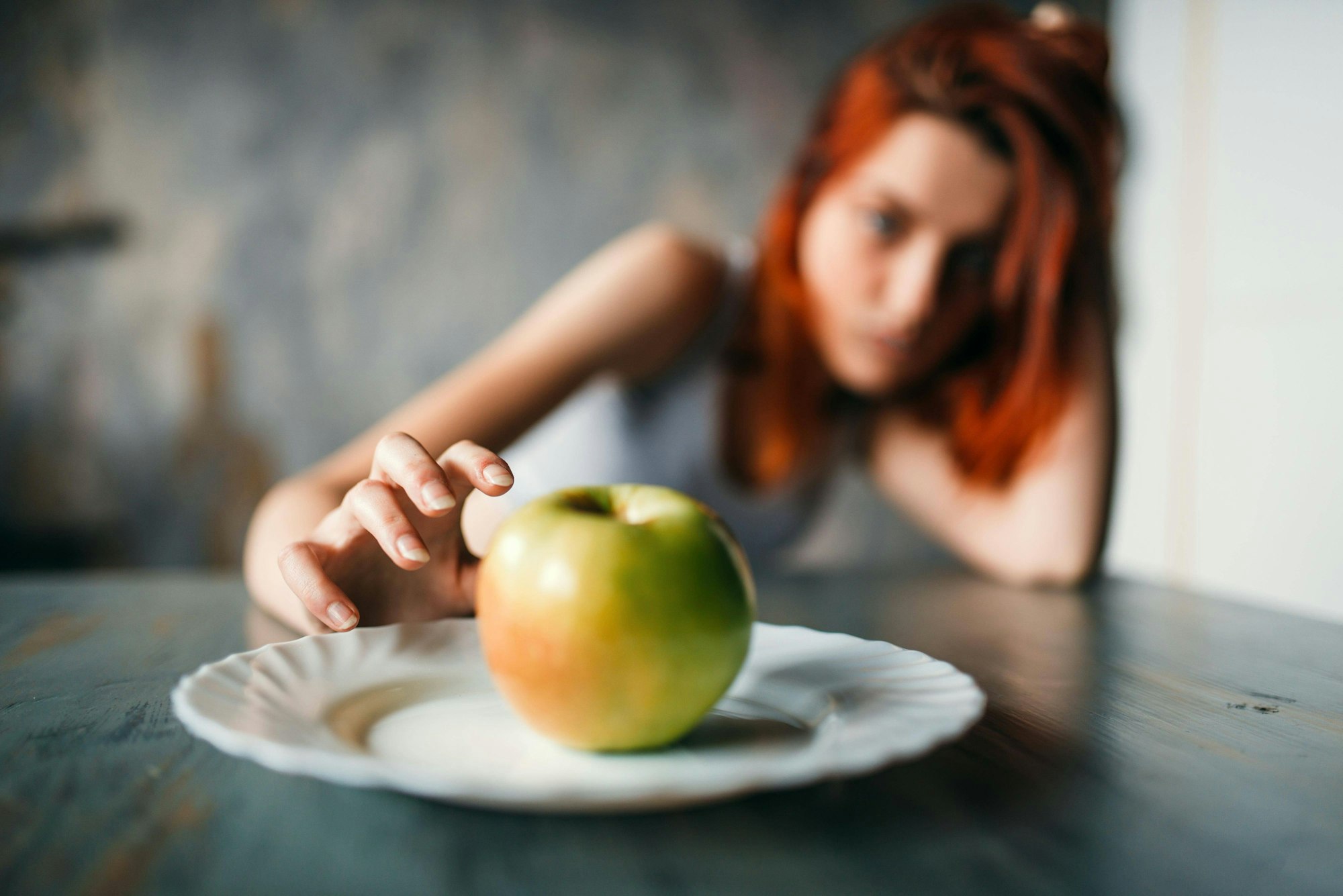 Eine dünne Jugendliche mit roten Haaren sitzt am Tisch, auf dem ein Teller mit einem Apfel steht.