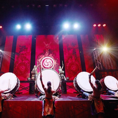 Fünf Trommler sitzen vor großen Trommeln, beugen sich mit freiem Oberkörper nach hinten, zum Schlag ansetzend.
