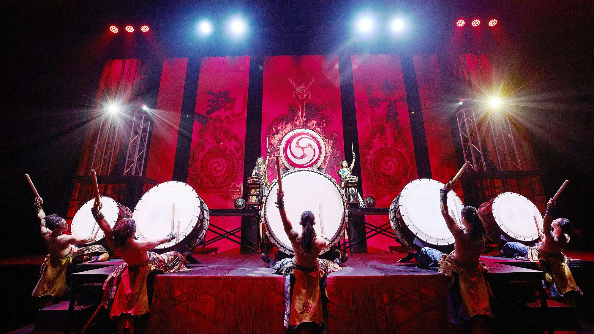 Fünf Trommler sitzen vor großen Trommeln, beugen sich mit freiem Oberkörper nach hinten, zum Schlag ansetzend.