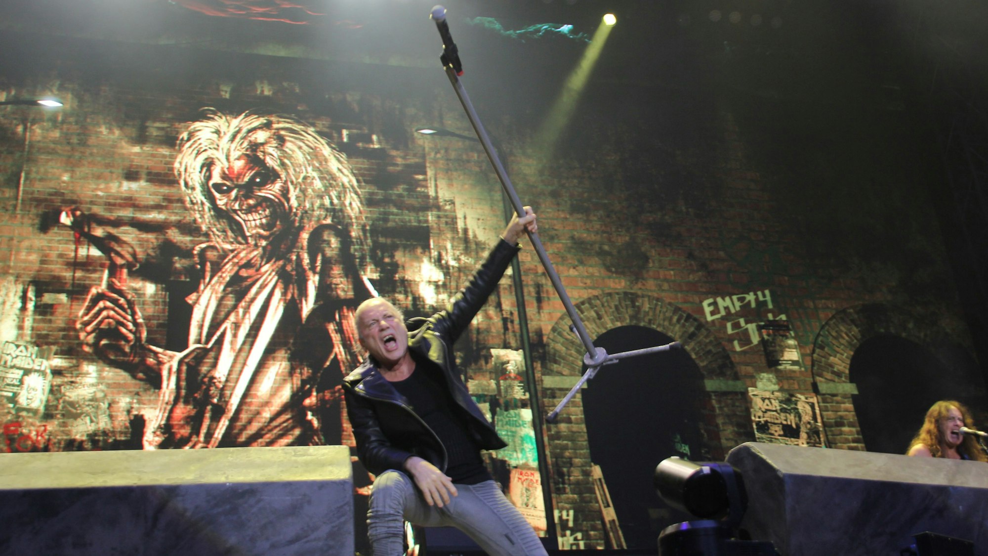 Iron-Maiden-Sänger Bruce Dickinson reckt seinen Mikrofonständer in die Höhe. Hinter ihm prangt Band-Maskottchen Eddie auf einem Videomonitor.