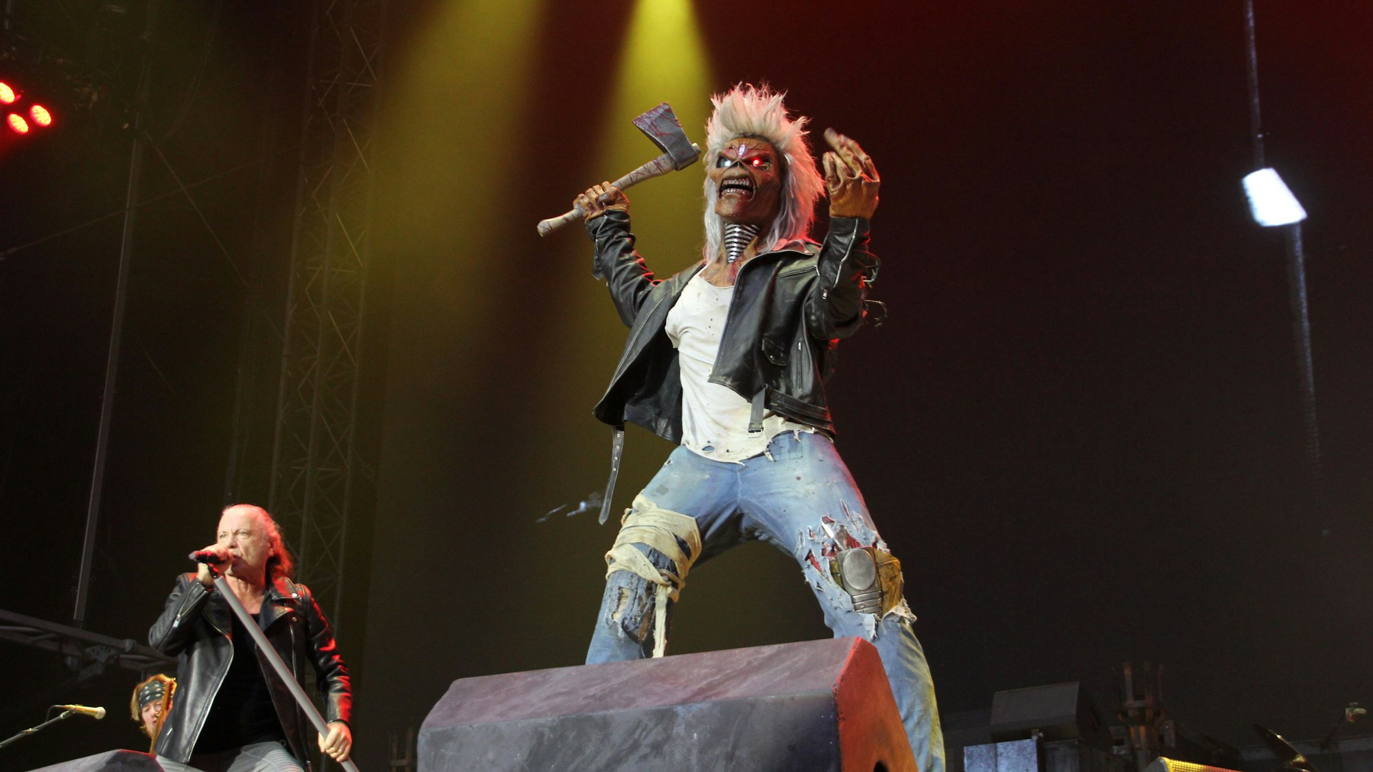Das Maskottchen der Band Iron Maiden ist ein überlebensgroßes Monster. Er steht auf der Bühne und schwingt bedrohlich eine blutgetränkte Axt. Rechts von Eddie steht Sänger Bruce Dickinson am Mikrofon.