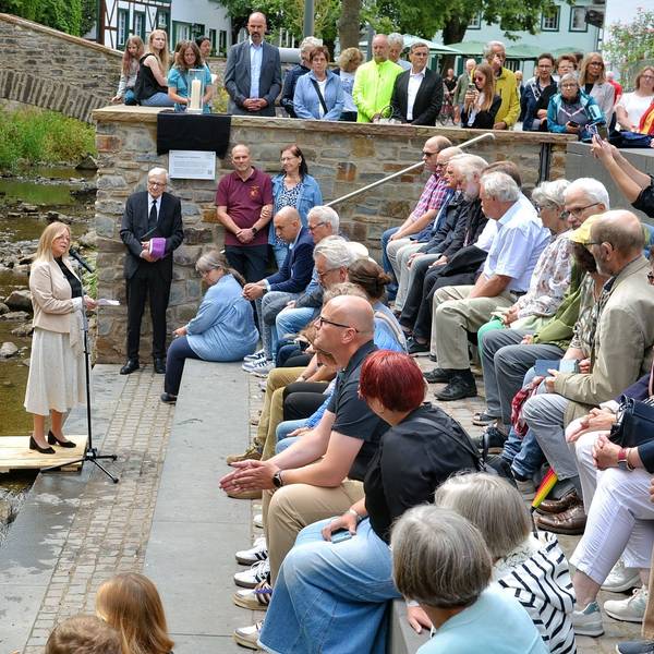 Ein Strauß Sonnenblumen steht in Bad Münstereifel in der Erft. Bürgermeisterin Sabine Preiser-Marian steht auf einem kleinen Podest und spricht in ein Mikrofon. Zahlreiche Menschen sitzen auf Steinstufen und hören ihr zu.