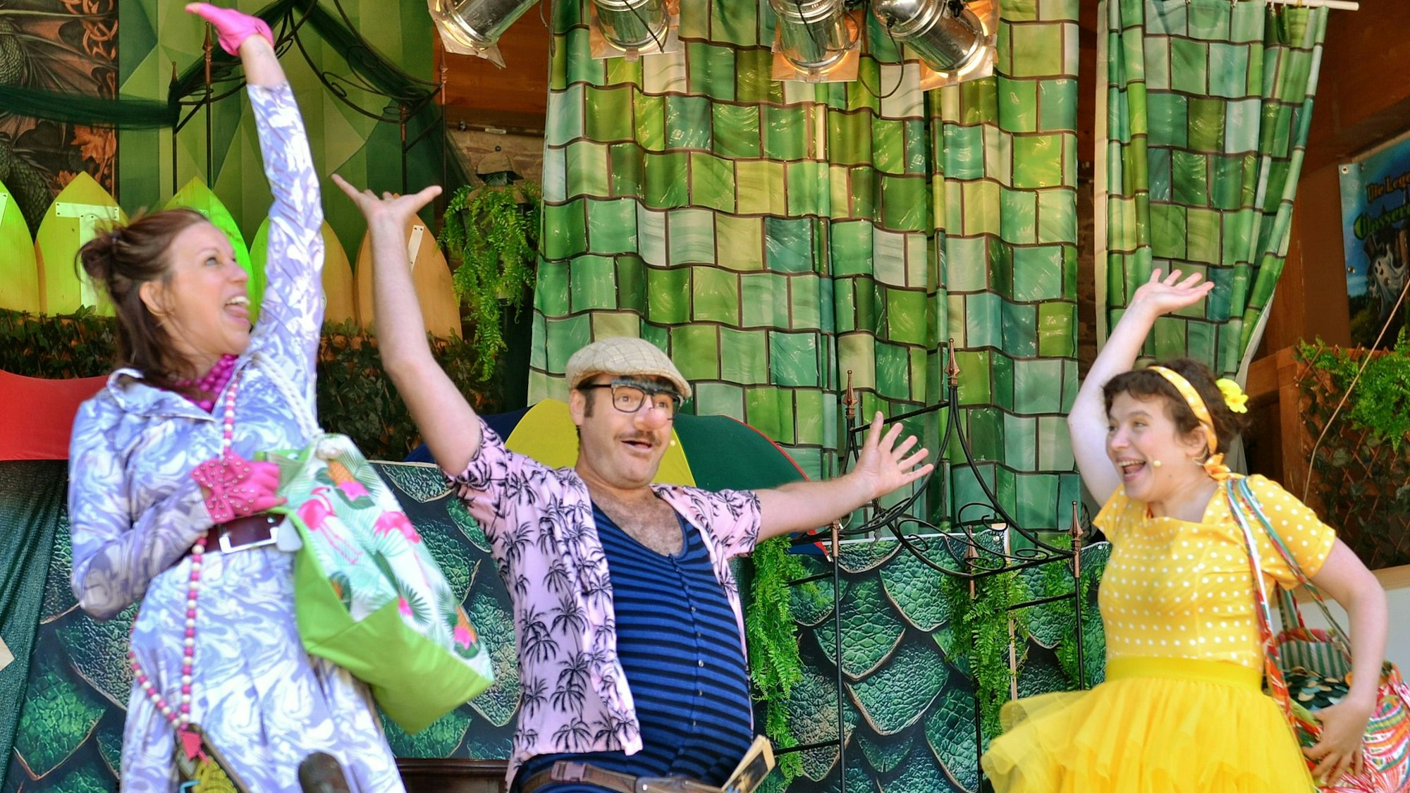 Zwei Frauen und ein Mann in bunter Strandkleidung auf einer Bühne lachen und heben die Arme in die Höhe.