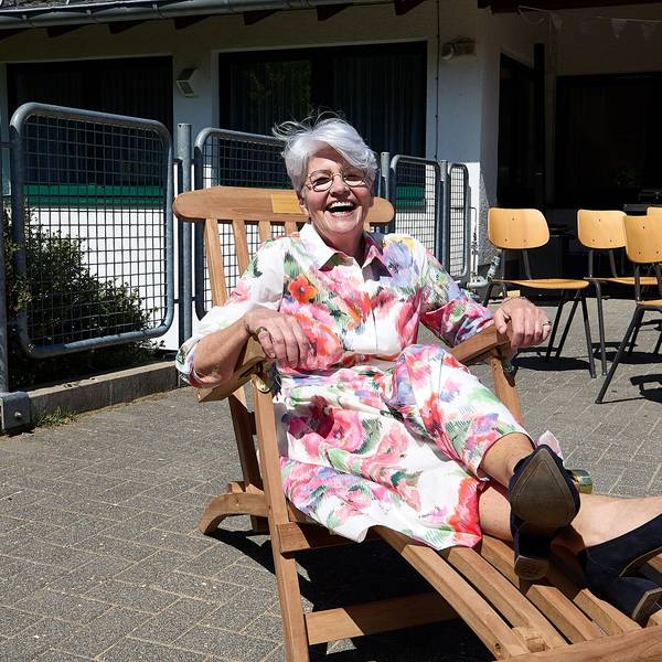 Das Bild zeigt eine Frau in einem bunten Kleid mit Blumenmotiven, die in einem Liegestuhl aus Holz sitzt und fröhlich lacht.