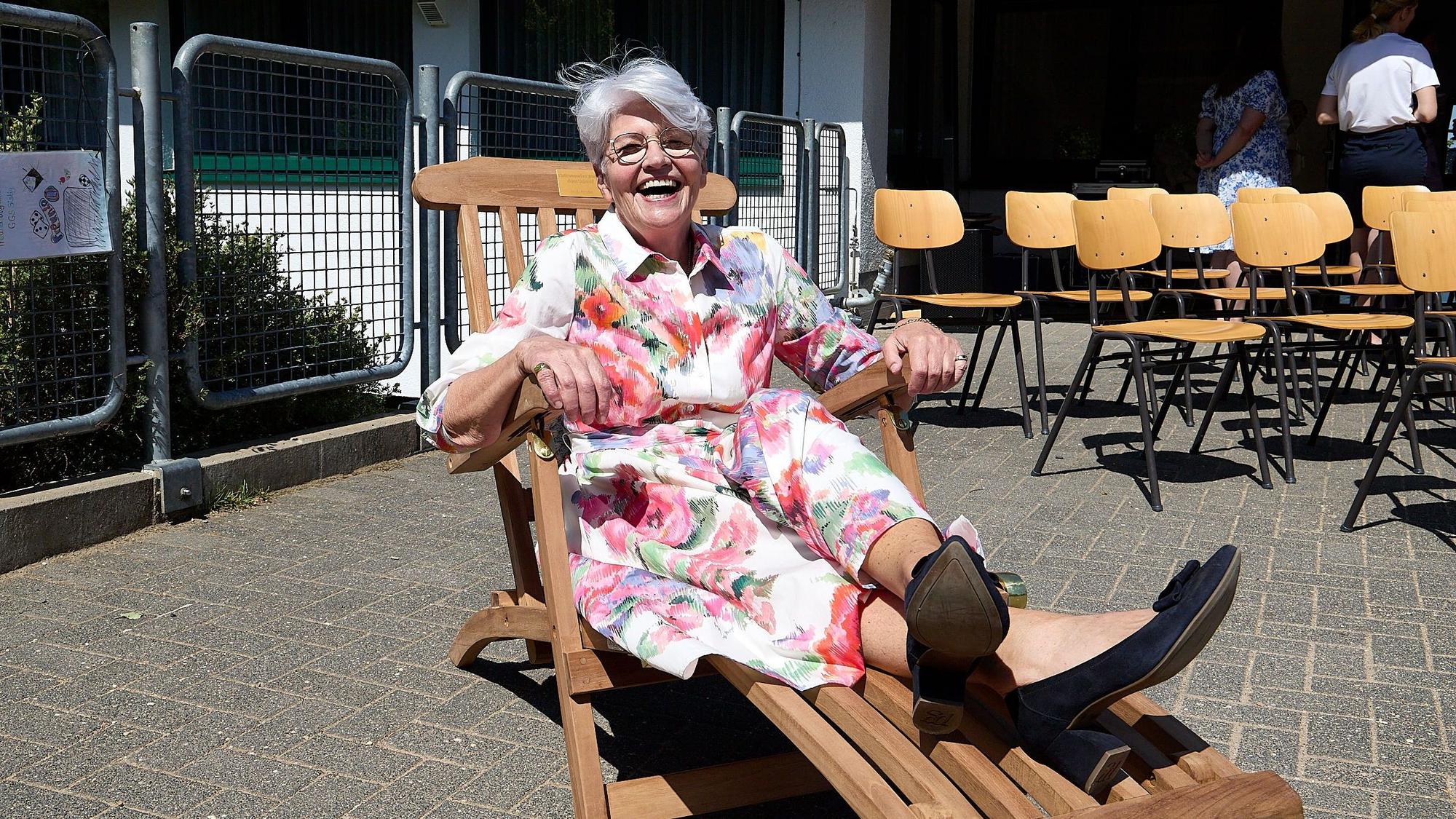 Das Bild zeigt eine Frau in einem bunten Kleid mit Blumenmotiven, die in einem Liegestuhl aus Holz sitzt und fröhlich lacht.