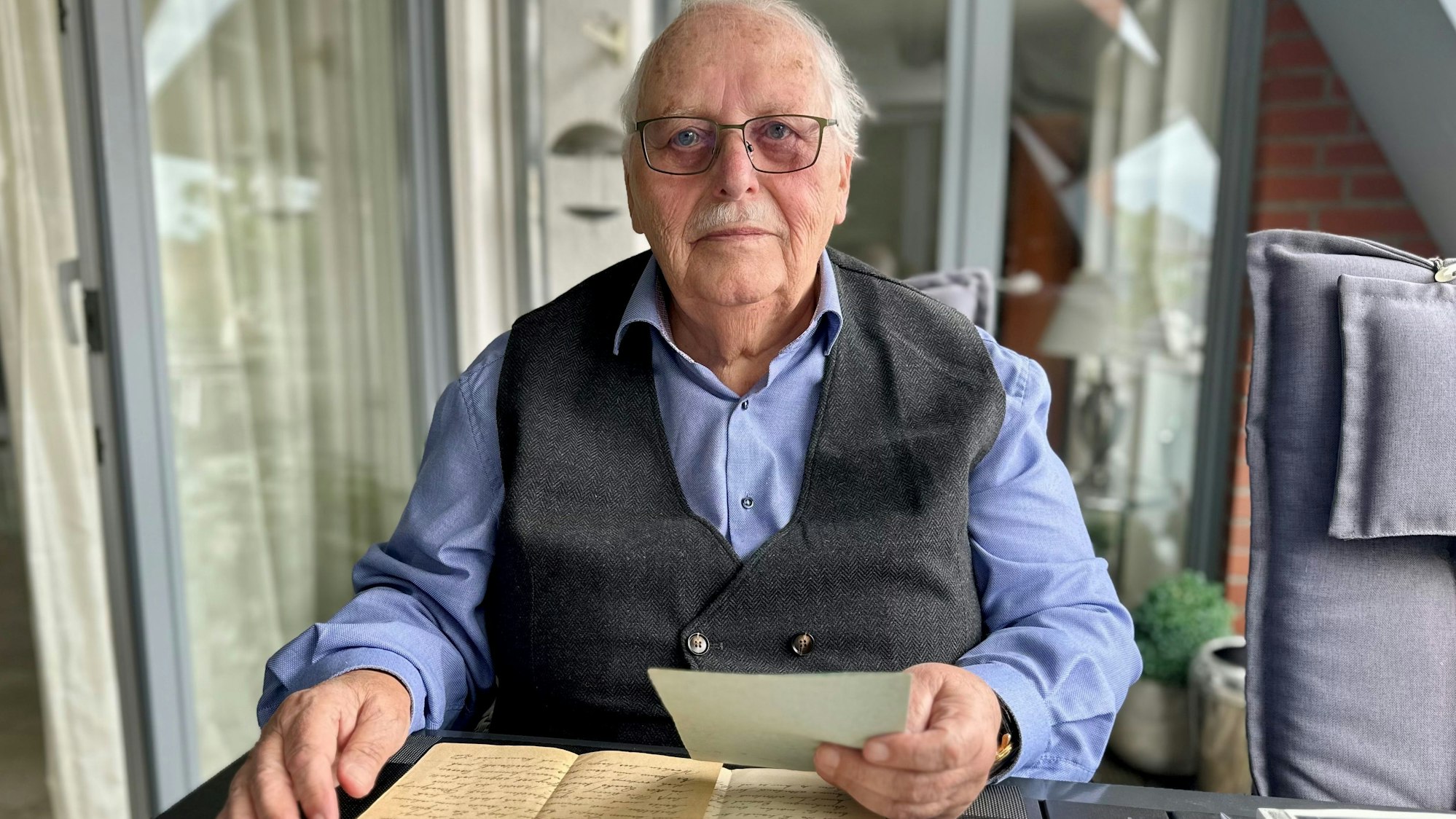 Zu sehen ist ein Mann, der einem Tisch sitzt und einen Brief in der Hand hält.