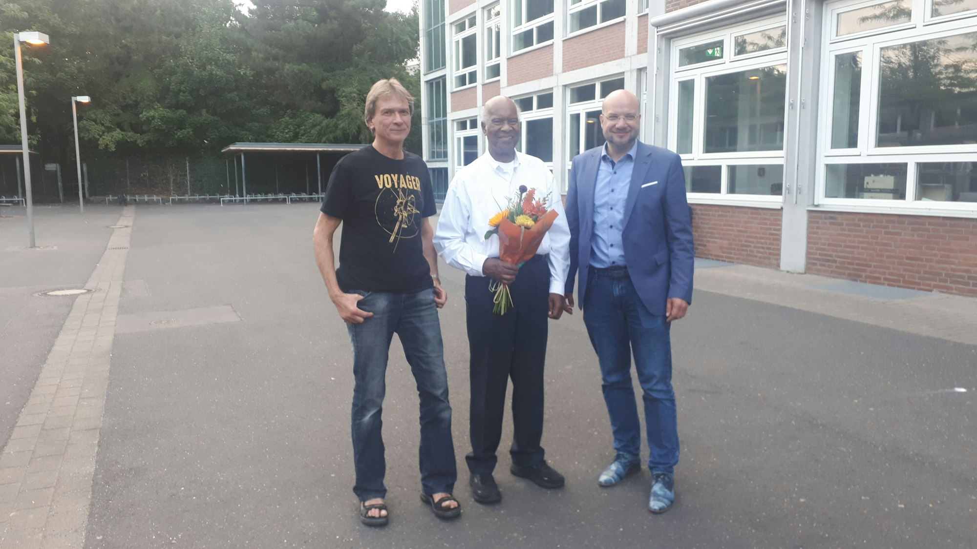 Drei Männer stehen auf einem Schulhof, der Mann in der Mitte hält einen Blumenstrauß in den Händen.