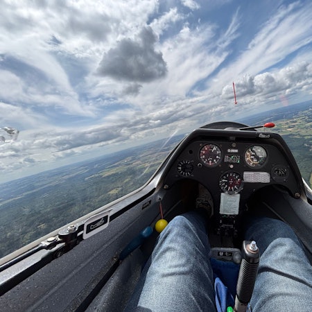 Foto aus einem Segelflugzeug. Der Blick geht aufs Instrumentenbrett, auf die Landschaft unten und die Wolken.
