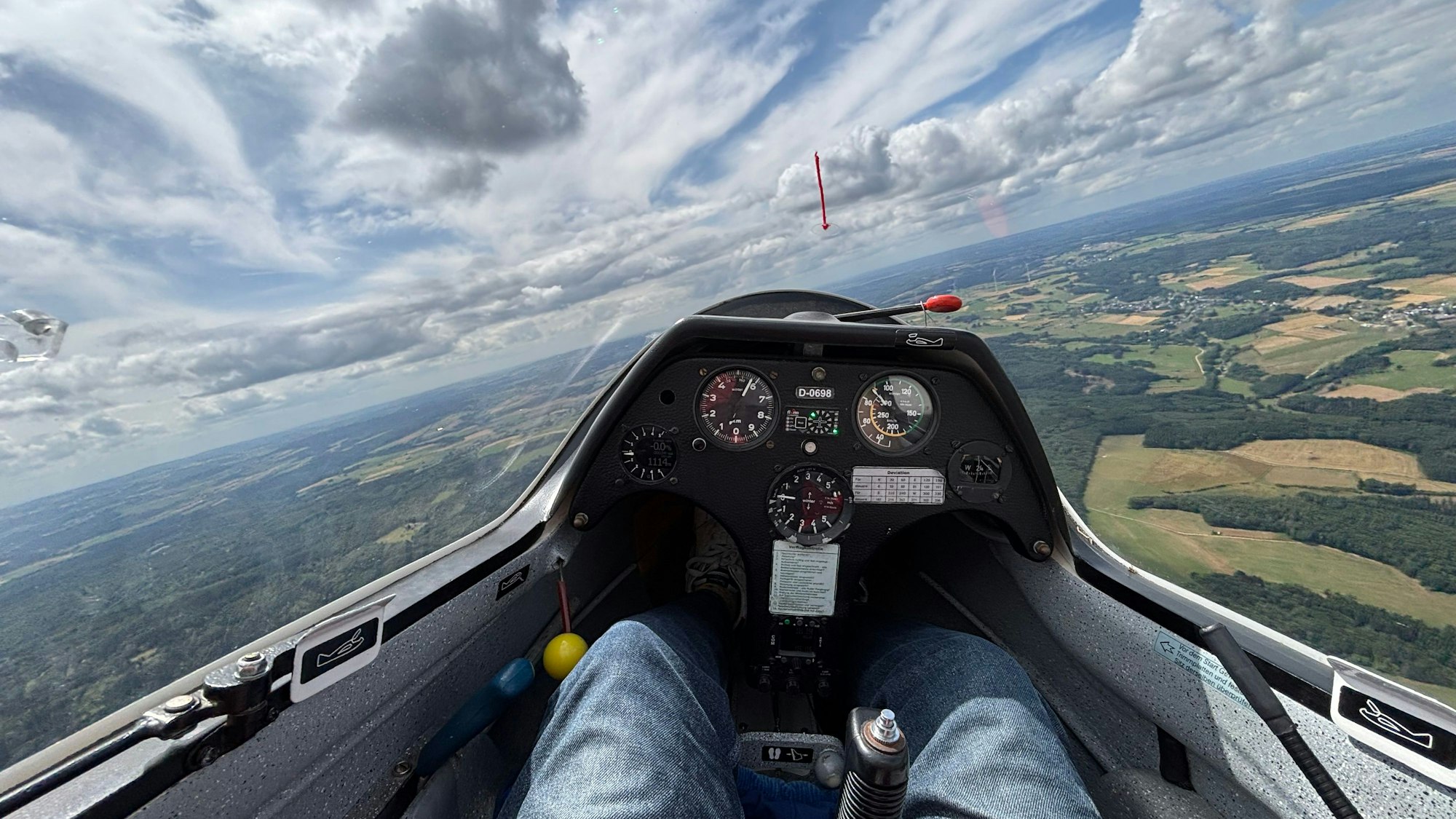 Foto aus einem Segelflugzeug. Der Blick geht aufs Instrumentenbrett, auf die Landschaft unten und die Wolken.
