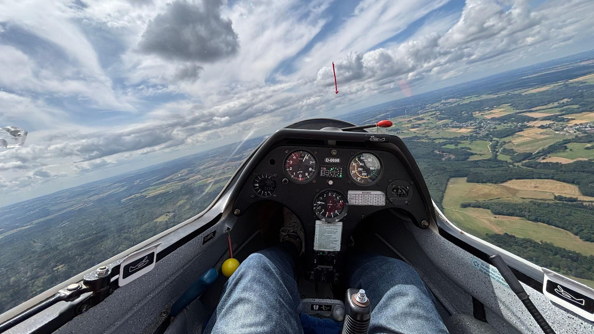 Foto aus einem Segelflugzeug. Der Blick geht aufs Instrumentenbrett, auf die Landschaft unten und die Wolken.