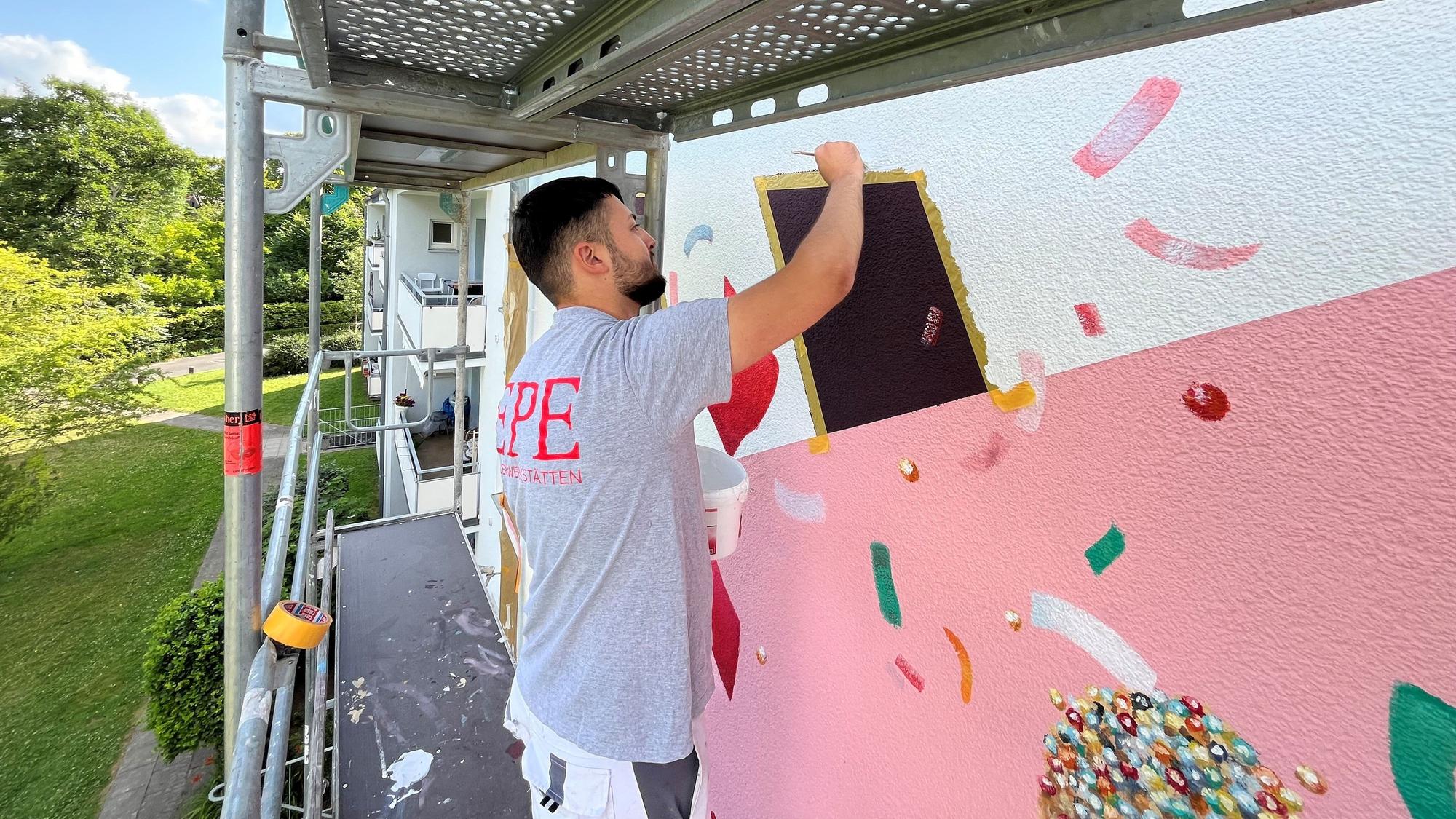 Ein Azubi der Epe Malerwerkstätten GmbH in Köln ist im Einsatz für ein Projekt gegen Vorurteile im Malerhandwerk und zeichnet an der kunstvollen Gestaltung einer Hausfassade in Porz.
