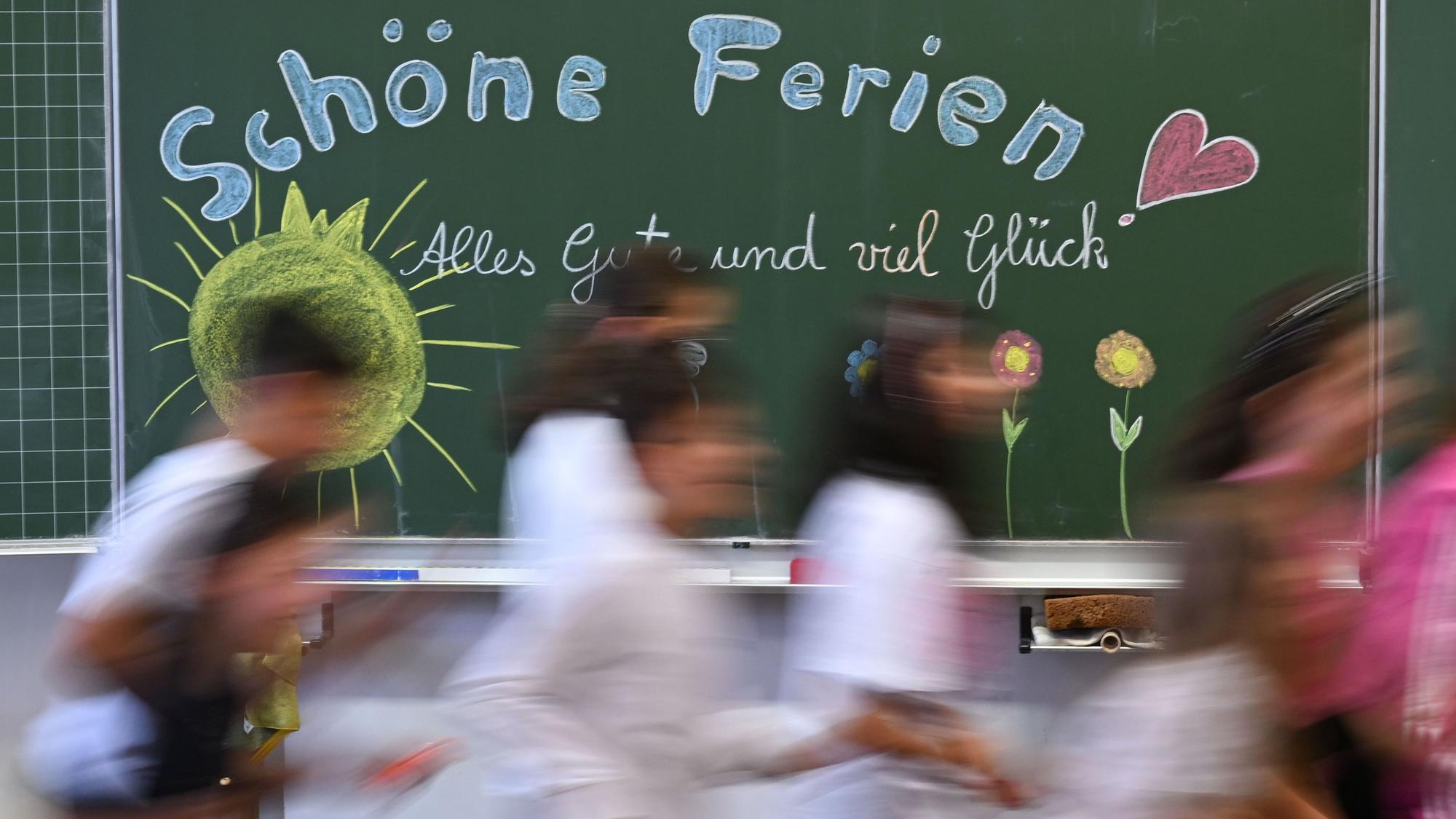 Schulkinder laufen am letzten Schultag an einer Tafel mit der Aufschrift „Schöne Ferien“ vorbei.