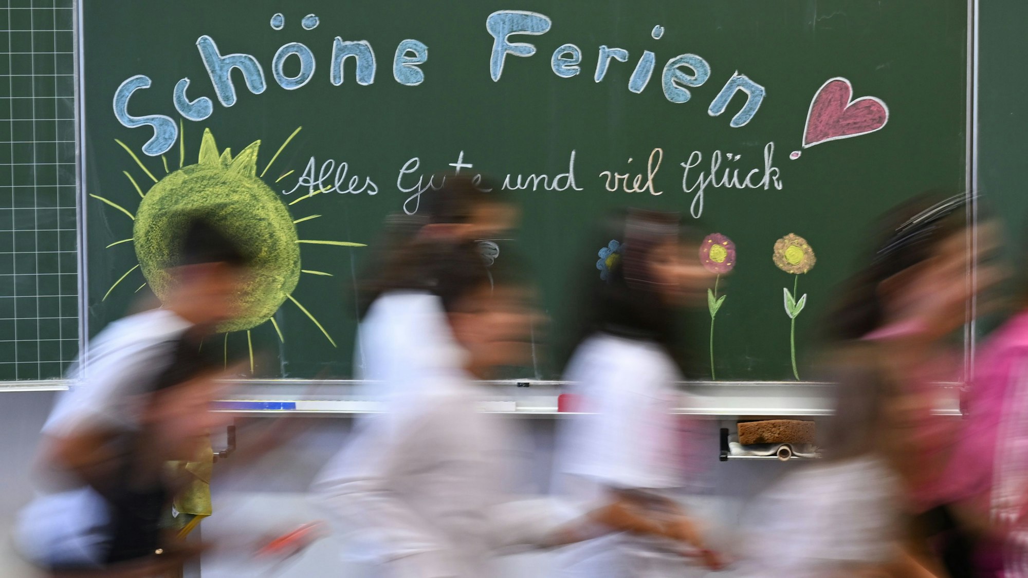 Schulkinder laufen am letzten Schultag an einer Tafel mit der Aufschrift „Schöne Ferien“ vorbei.