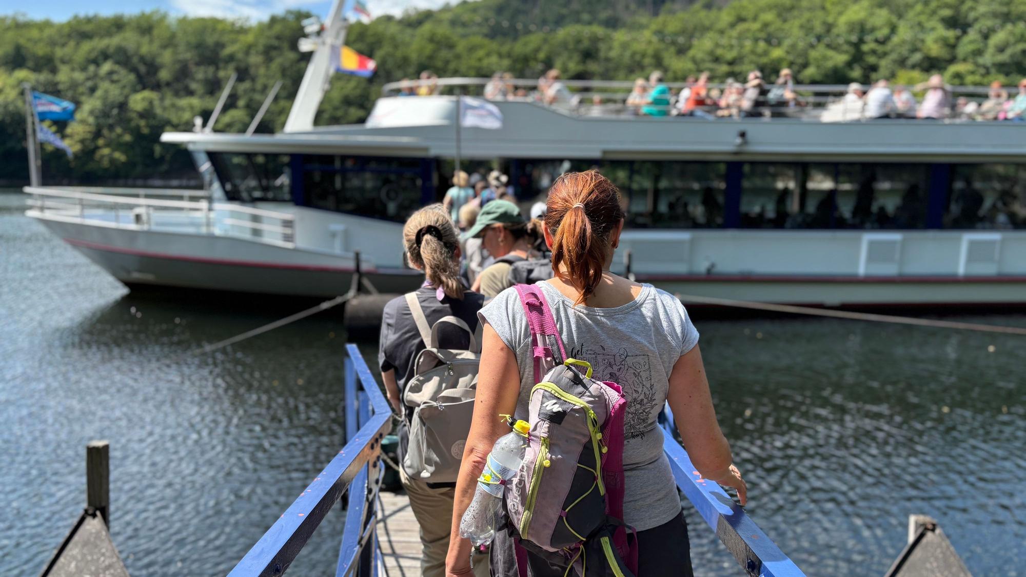 Die Wandergruppe geht mit dem Rücken zur Kamera einen Steg in Richtung eines Schiffs entlang.