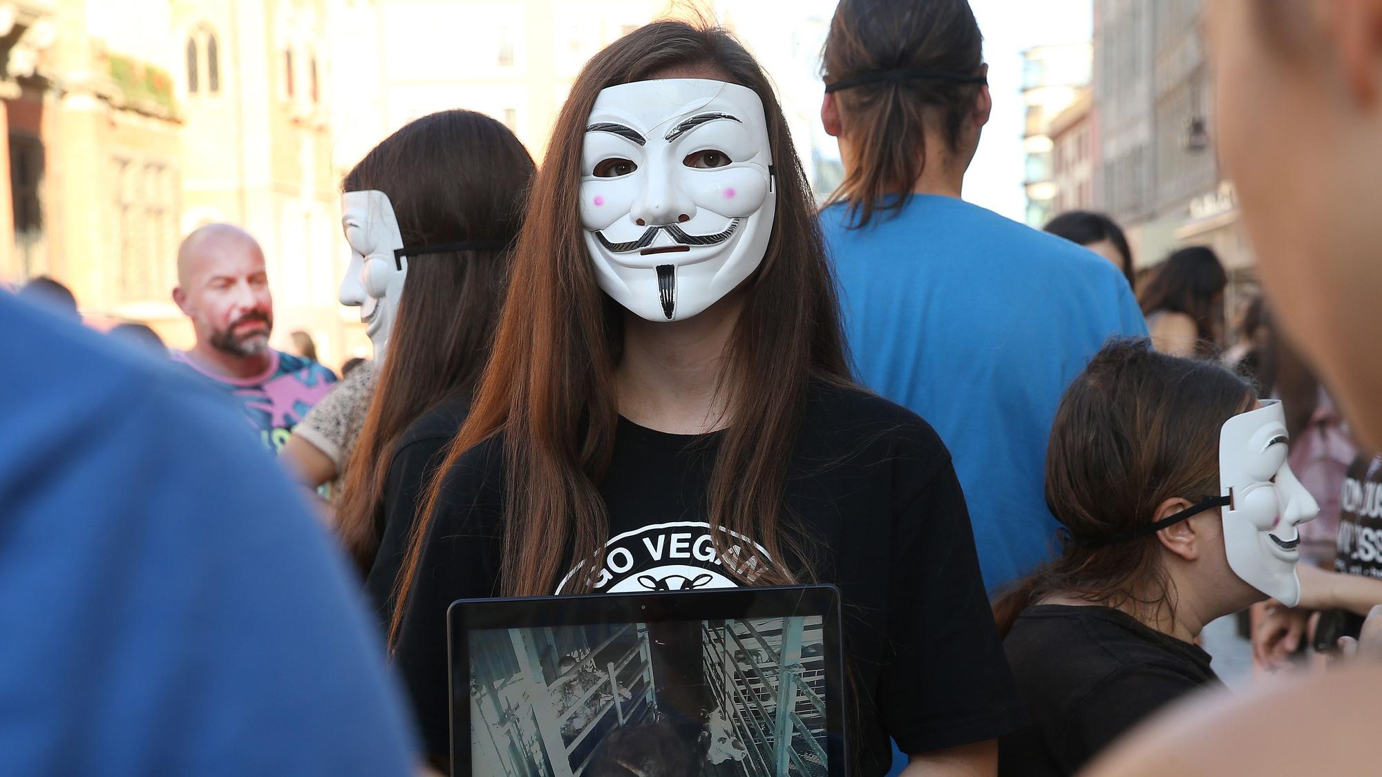 Eine Person von Anonymous for the Voiceless steht mit einem Bildschirm und einer Maske auf einem Platz in Polen. Um sie herum stehen weitere Menschen.