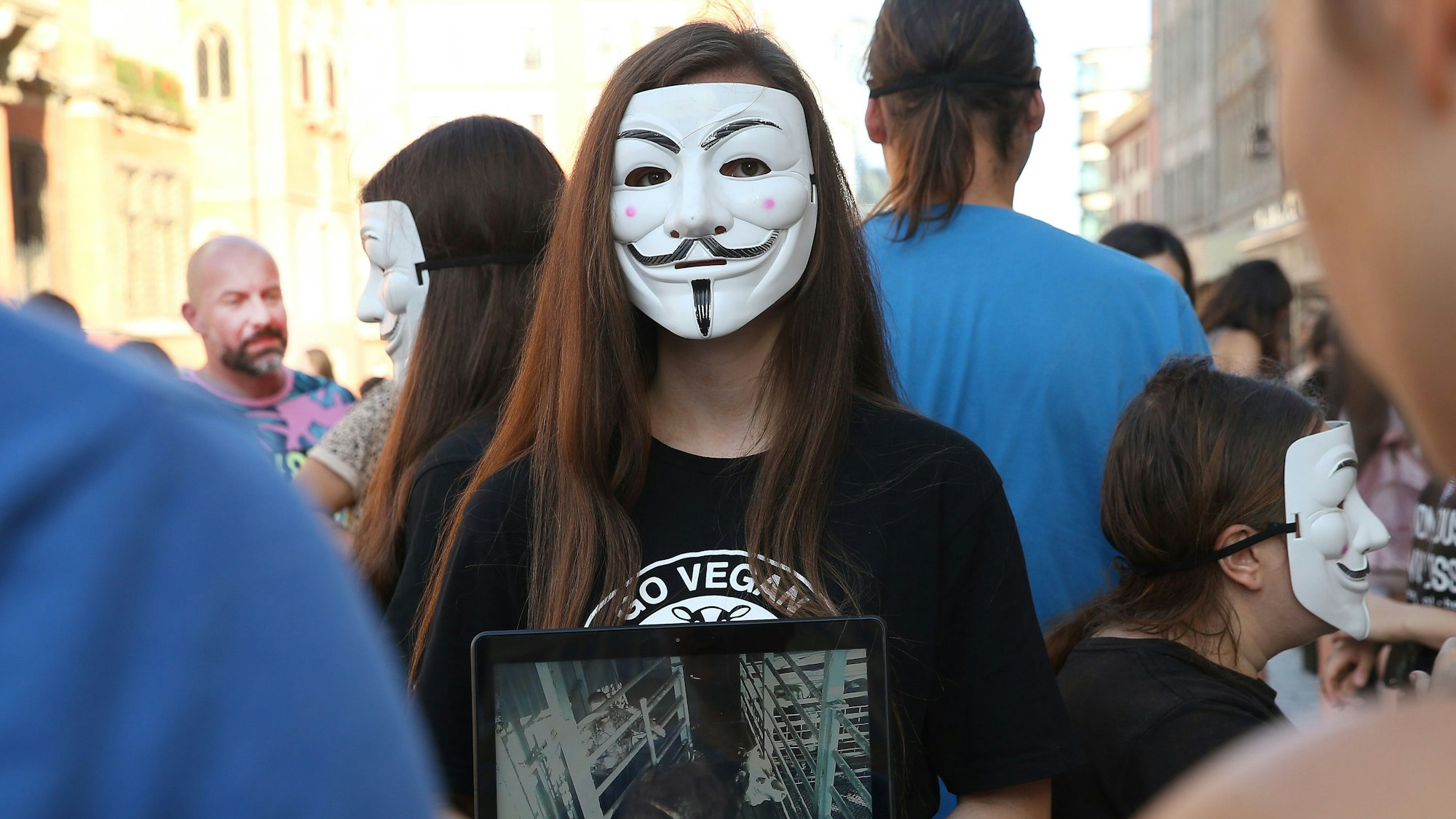 Eine Person von Anonymous for the Voiceless steht mit einem Bildschirm und einer Maske auf einem Platz in Polen. Um sie herum stehen weitere Menschen.