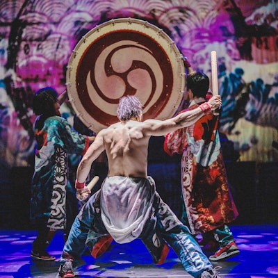 Ein Trommler mit bloßem Oberkörper macht sich zum Schlag auf eine große Trommel bereit, die von zwei anderen Männern hochgehalten wird.