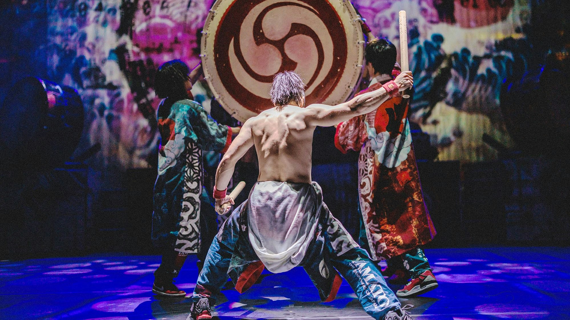 Ein Trommler mit bloßem Oberkörper macht sich zum Schlag auf eine große Trommel bereit, die von zwei anderen Männern hochgehalten wird.