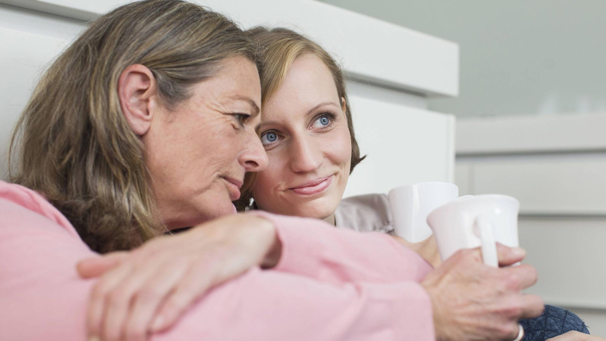 Eine ältere Frau und eine Teenagerin sitzen vor einer Wand und haben jeweils eine Tasse in der Hand.