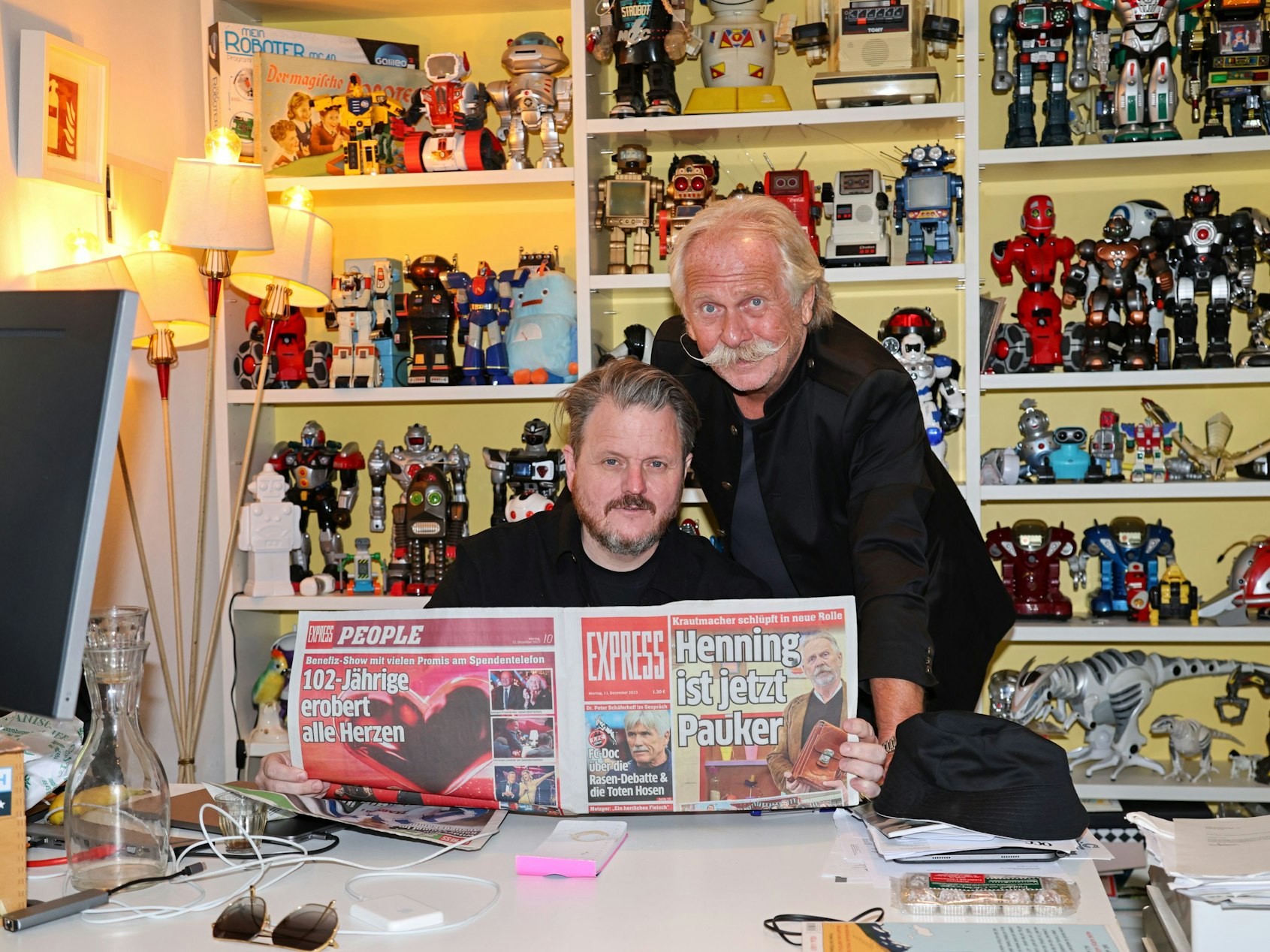 Zwei Männer sitzen vor einem aufgeschlagenen EXPRESS mit der Schlagzeile „Henning ist jetzt Pauker“.