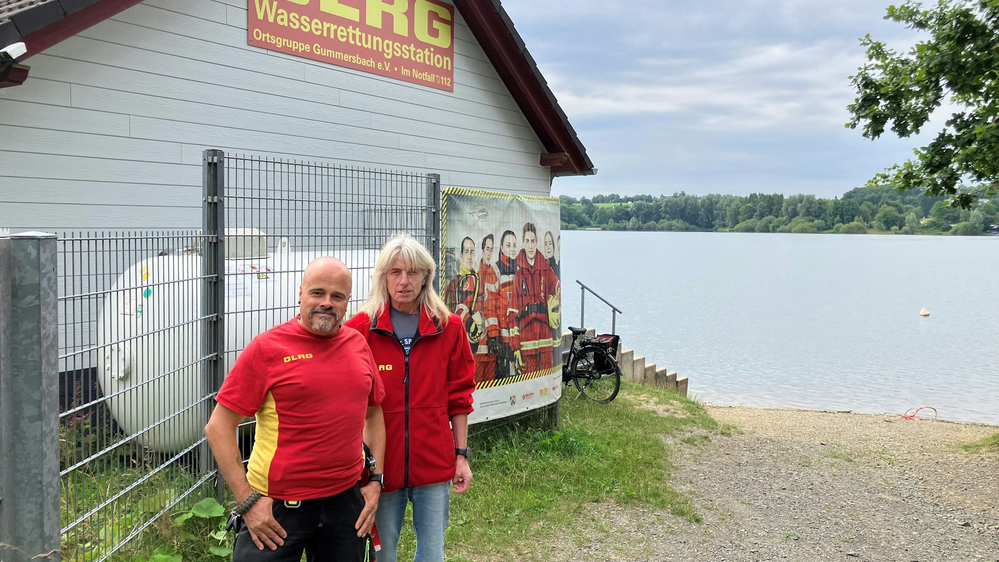 Ein Mann und eine Frau stehen vor einer Wachstation der DLRG an einem Badesee.