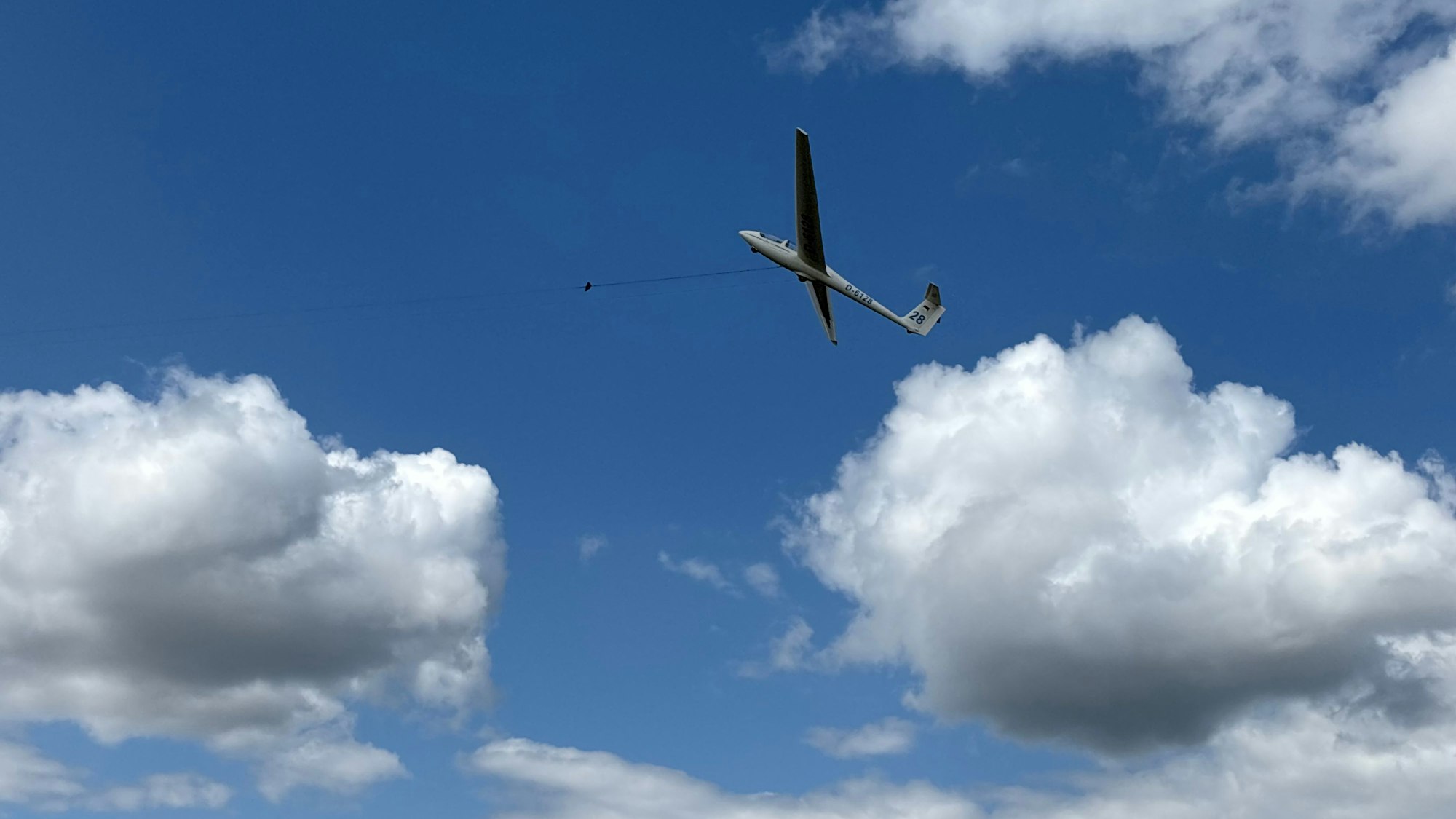 Ein Segelflugzeug, das an einem Seil hängt, befindet sich schräg in der Luft. Im Hintergrund sind Wolken.