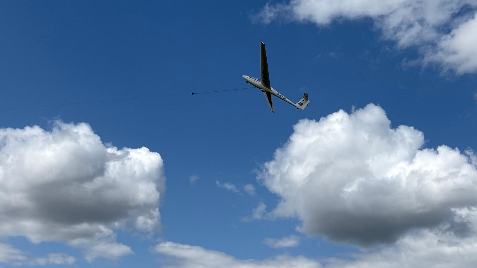 Ein Segelflugzeug, das an einem Seil hängt, befindet sich schräg in der Luft. Im Hintergrund sind Wolken.