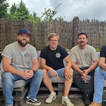Vier Männer in den 30ern sitzen nebeneinander auf einem Outdoor-Sofa.