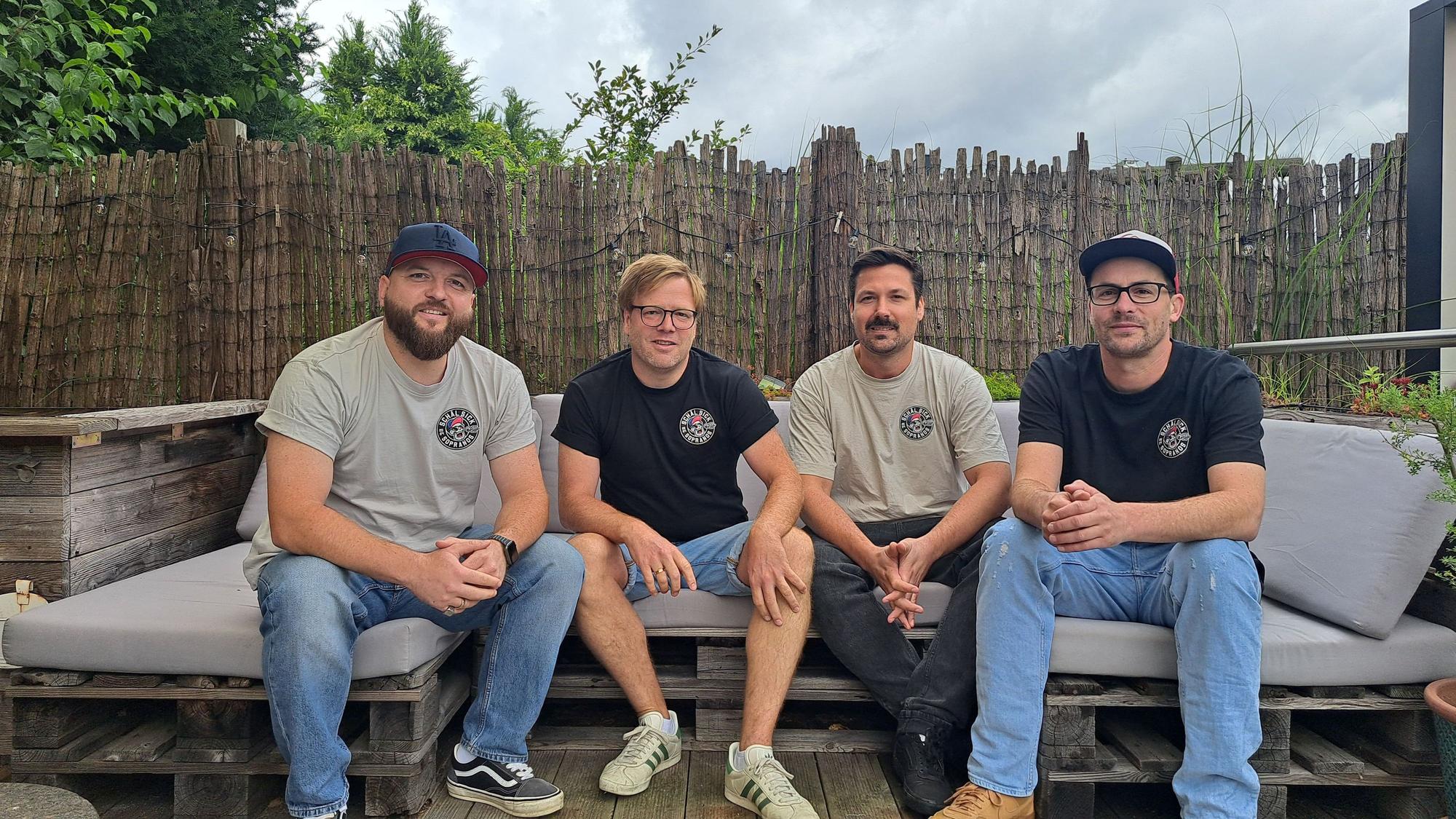 Vier Männer in den 30ern sitzen nebeneinander auf einem Outdoor-Sofa.