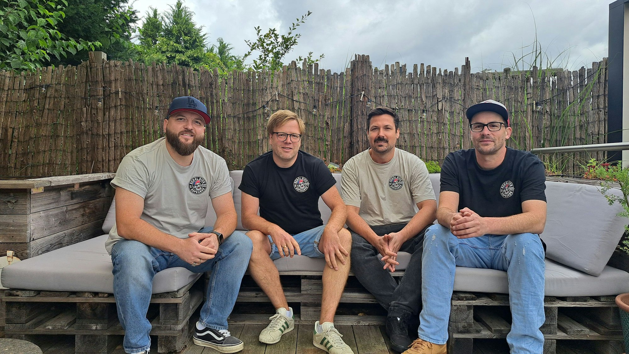 Vier Männer in den 30ern sitzen nebeneinander auf einem Outdoor-Sofa.
