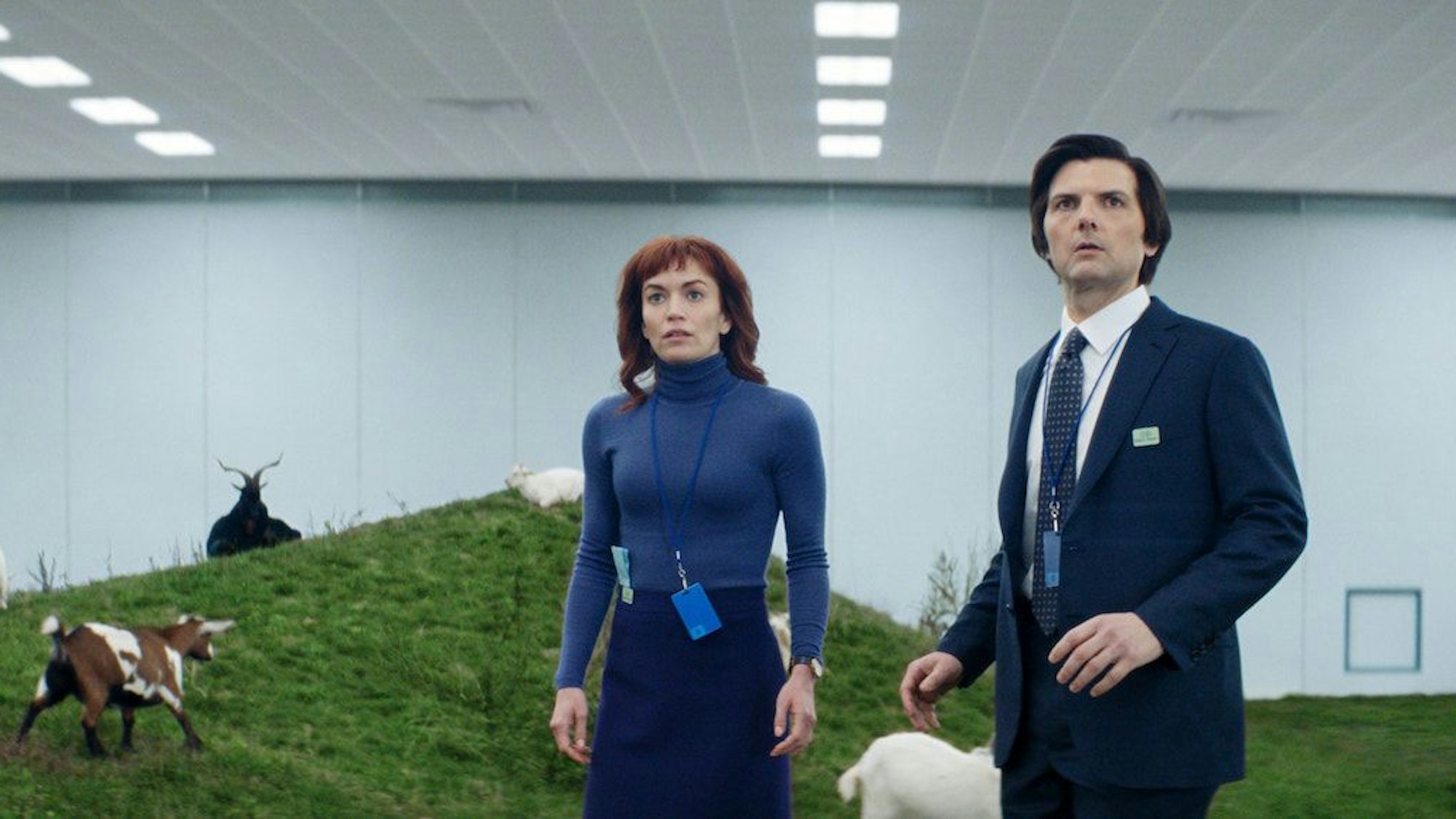 Britt Lower und Adam Scott stehen vor einer hügeligen Wiese mit Ziegen, die sich in einem riesigen weißen Büroraum befindet.