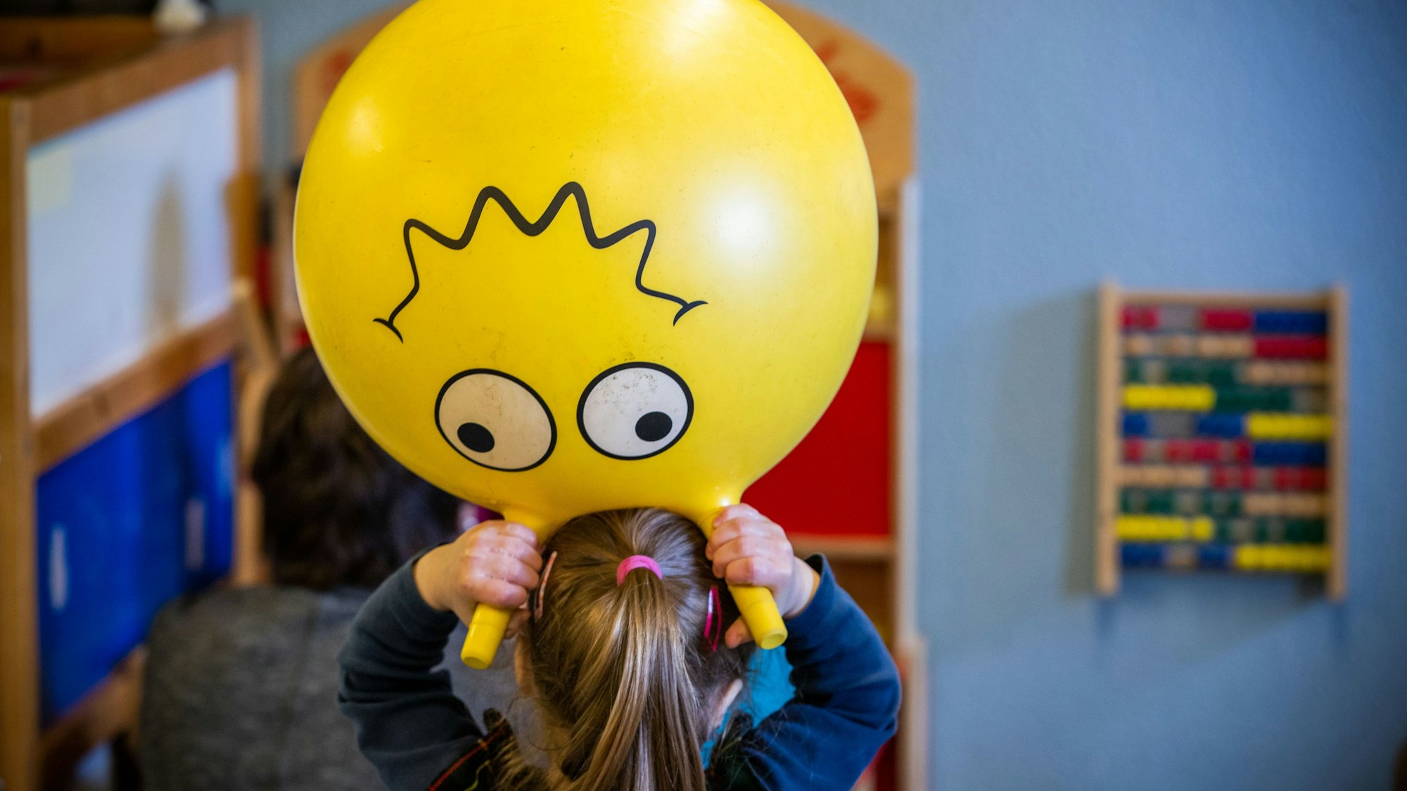 Ein Kind in einer Kindertagespflegeeinrichtung hält sich einen gelben Hüpfball mit aufgemalten Augen und Mund über den Kopf.