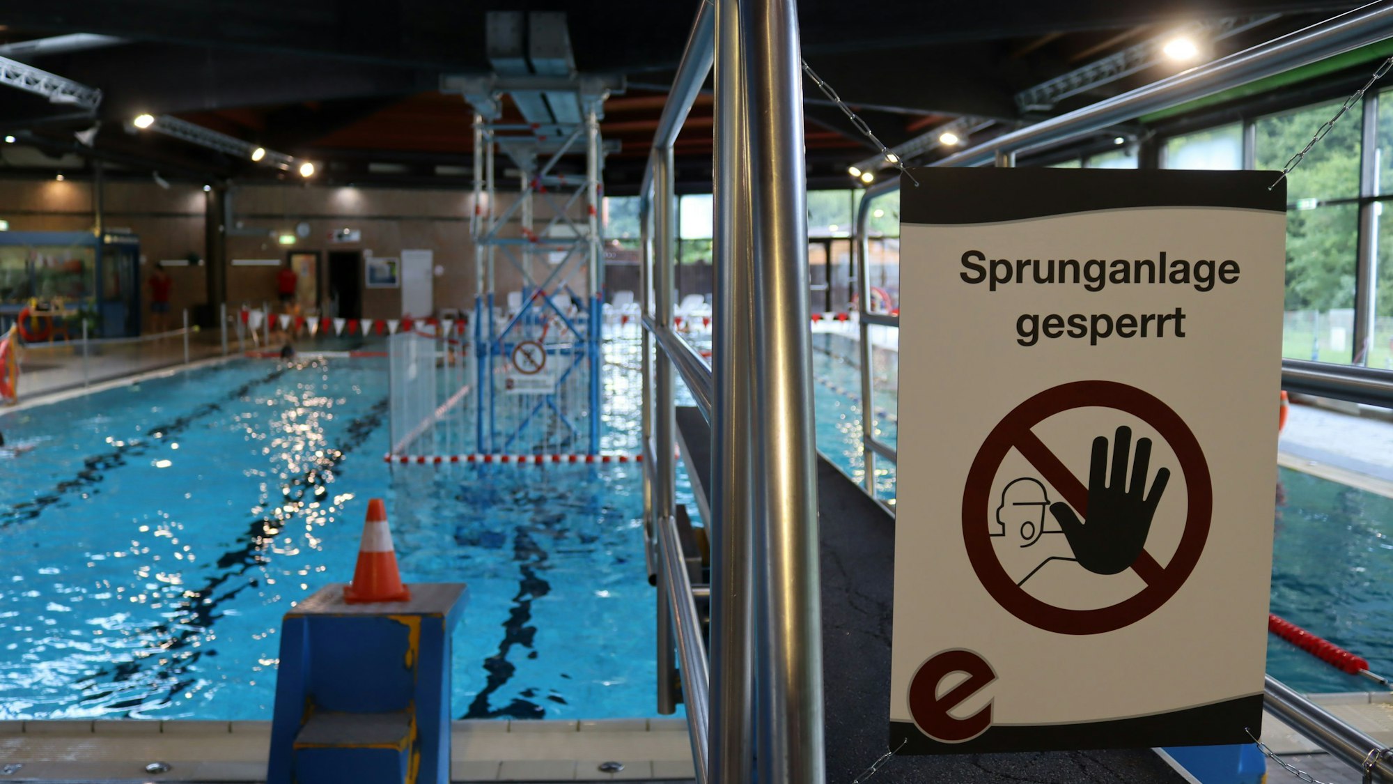 Ein Schild weist du den gesperrten Sprungturm hin. Orange-weiße Kegel auf den Böckchen, von den die Farbe abgeplatzt ist, deuten ebenfalls eine Sperrung an. Grund ist die Stützkonstruktion im Hintergrund, mitten im Schwimmerbecken.