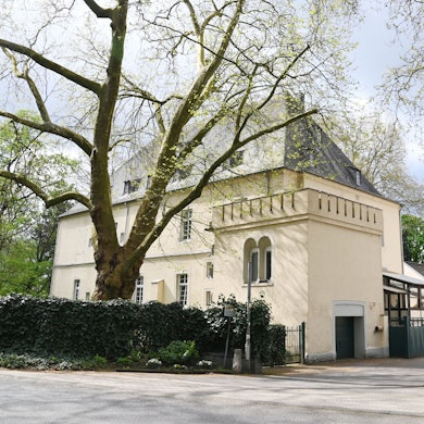 Ein großes herrschaftliches Gebäude mit Nebengebäuden und einer vergitterten Hofeinfahrt ist zu sehen.