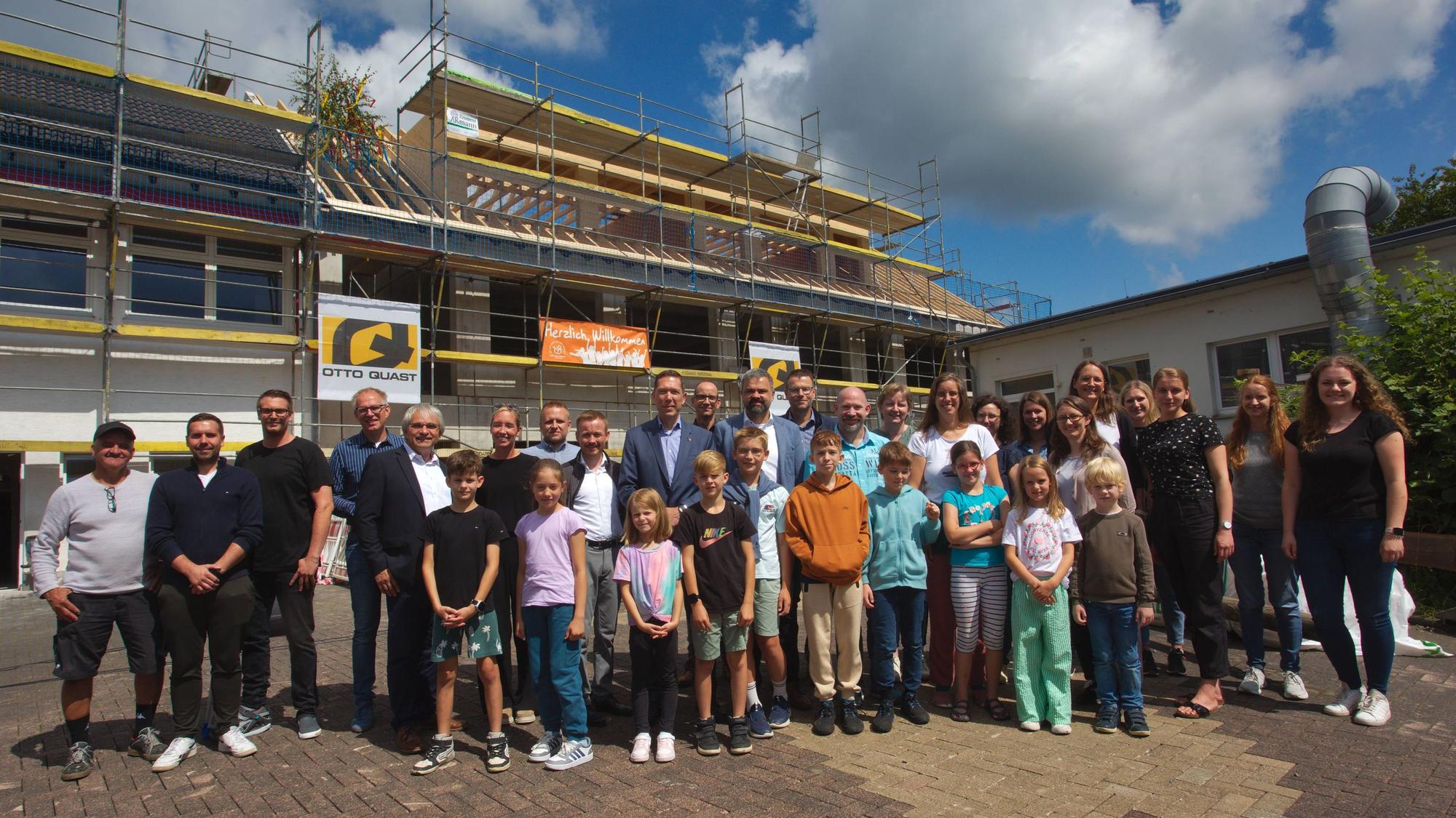 Verantwortliche, Eltern und Kinder stehen vor der Baustelle, mit dem neu gerichteten Dachstuhl des Anbaus.