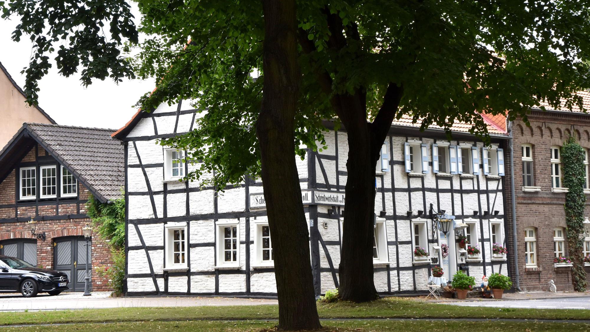 Zu sehen ist ein weißes Haus mit schwarzem Fachwerk hinter zwei großen Bäumen.