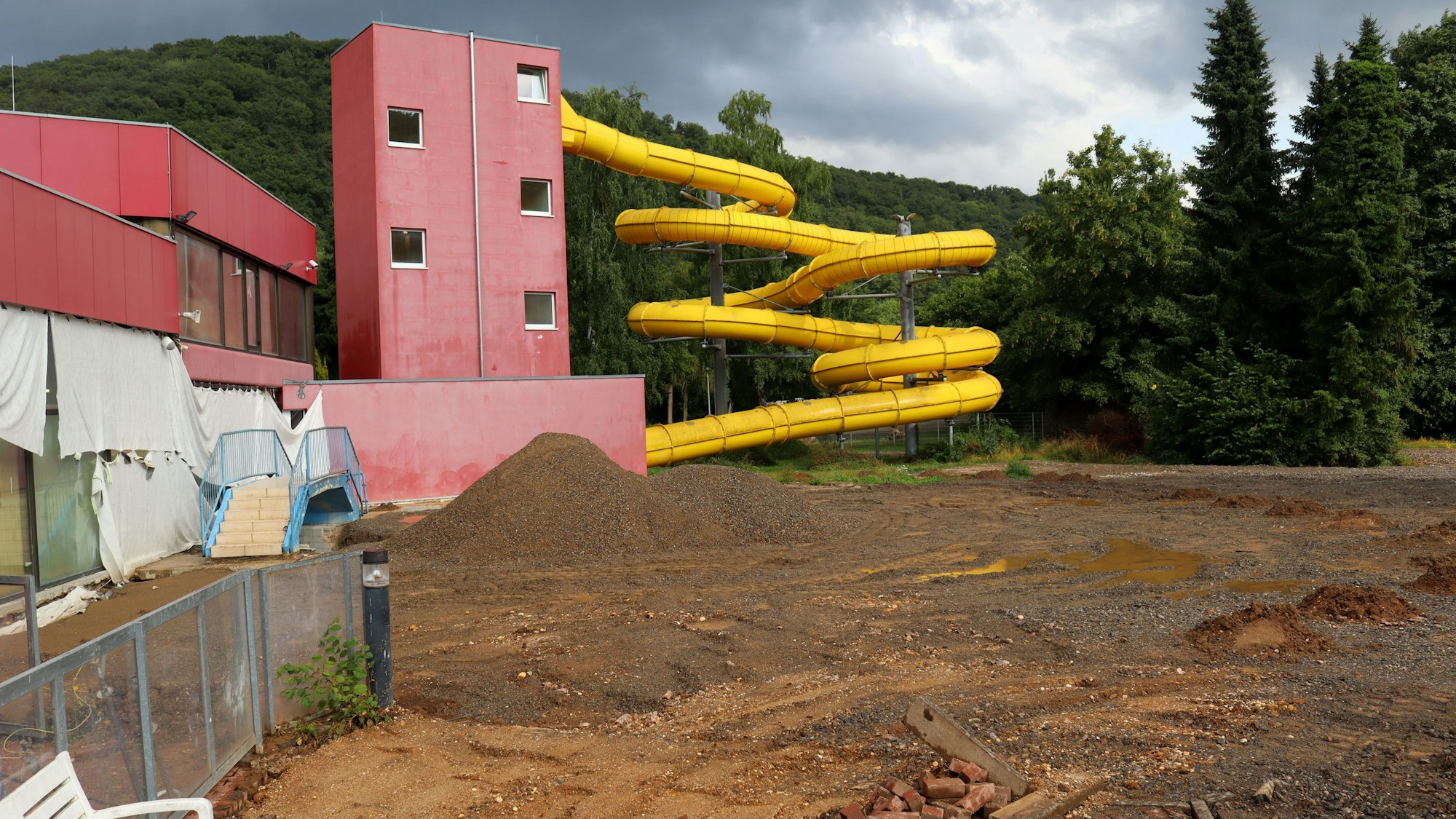 Eine Erdfläche ist zu sehen. Dort kommt das Außenbecken des Eifelbades hin. Im Hintergrund die Rutsche des Schwimmbades.
