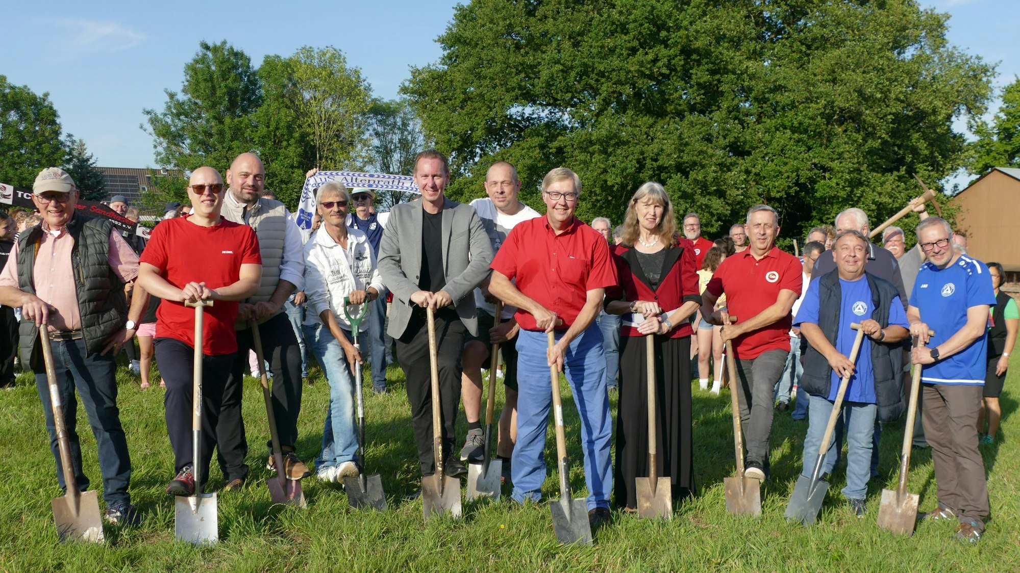 Männer und Frauen auf einer Wiese; sie halten Spaten vor sich.