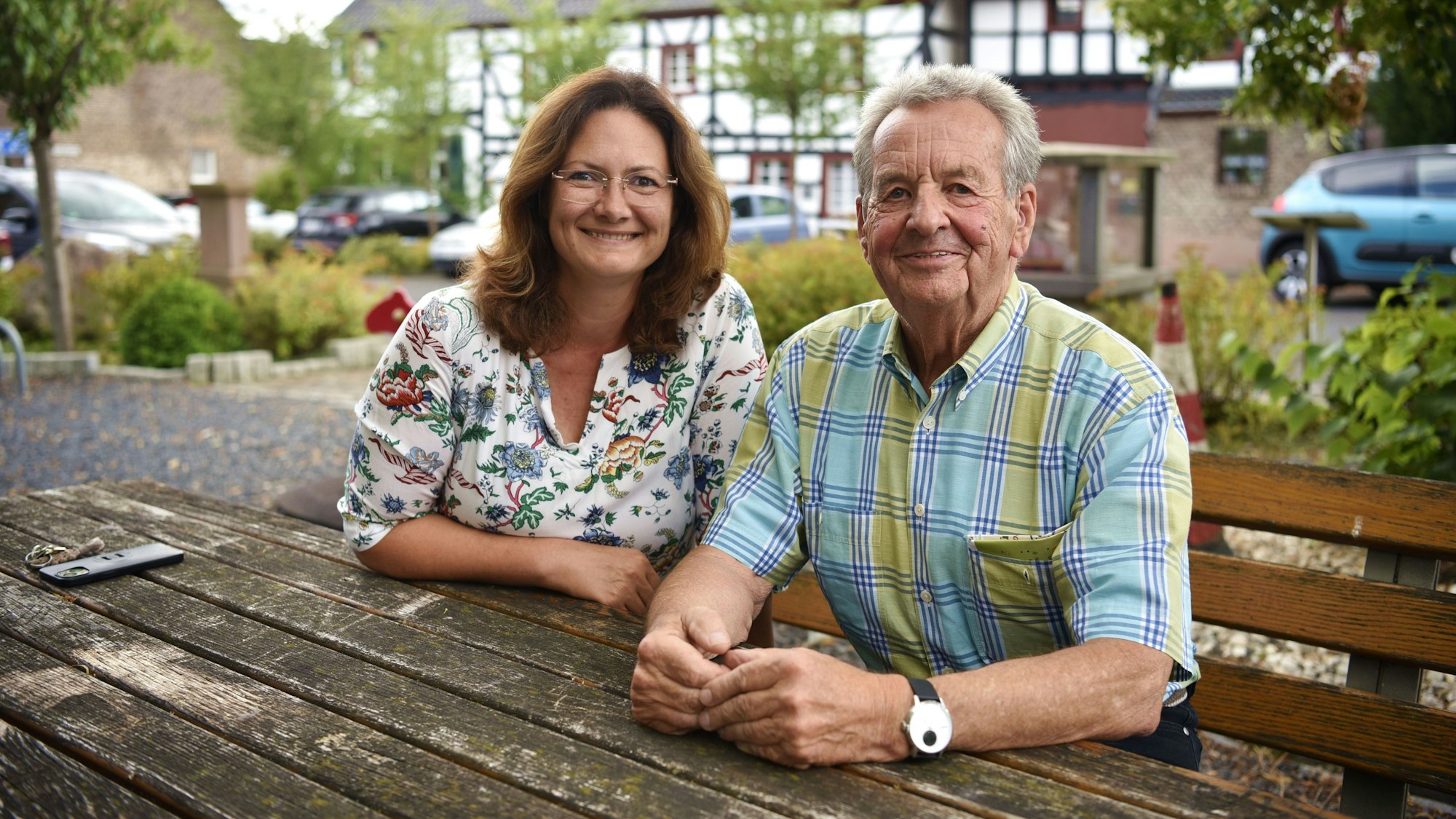 Tamara Porschen und Josef Heinrichs von „Zukunft für Sinzenich“ sitzen auf einer Bank an einem Tisch auf dem Dorfplatz. Im Hintergrund sind Bäume und Fachwerkhäuser zu sehen.