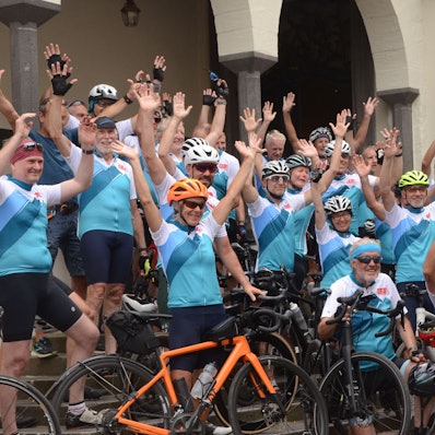 Männer und Frauen in Rennradtrikots werfen die Hände in die Luft.