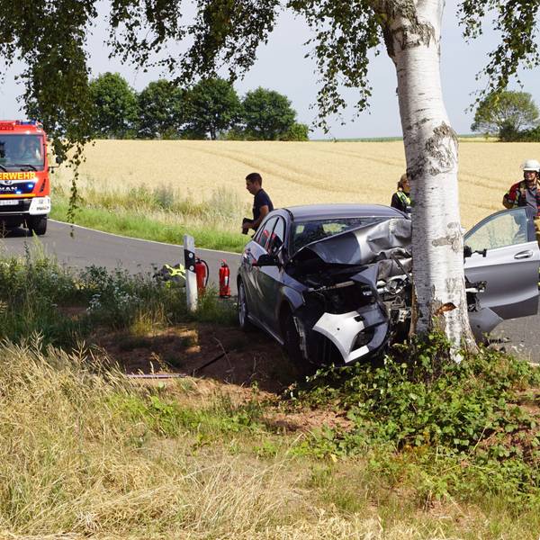 Das Foto zeigt den zerstörten Pkw am Baum, daneben Polizisten und Feuerwehrleute und im Hintergrund auf der Straße ein Feuerwehrfahrzeug.