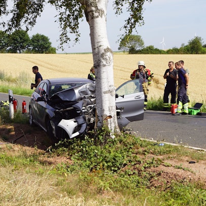 Das Foto zeigt den zerstörten Pkw am Baum, daneben Polizisten und Feuerwehrleute und im Hintergrund auf der Straße ein Feuerwehrfahrzeug.