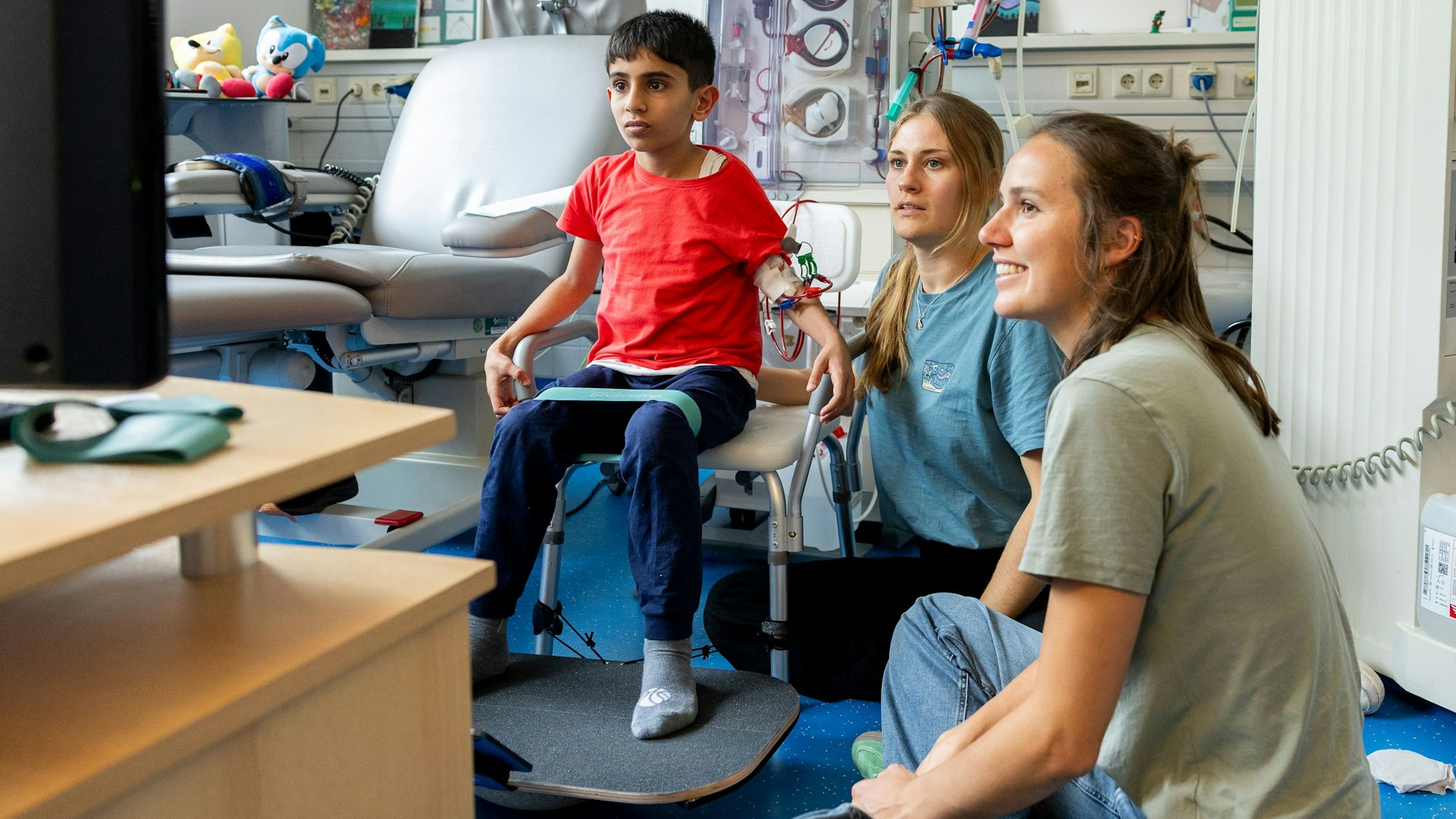 Mit seinen Füßen steuert Dialysepatient Goran ein Computerspiel, Marleen Kerstin (Mitte) und Lene Stegelmann (r.) von der Sporthochschule feuern ihn an und achten auf die richtige Körperhaltung.