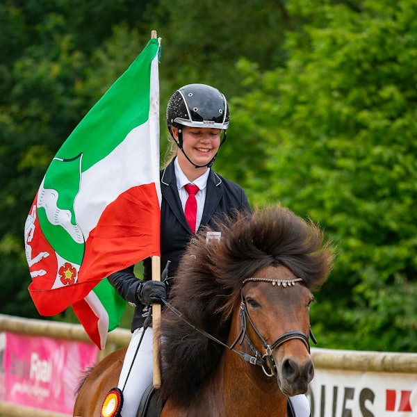 Emma Brust auf dem Rücken eines Pferdes. In der Hand hält sie eine NRW-Flagge.
