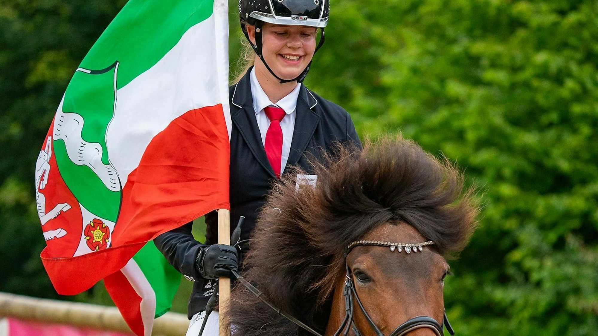 Emma Brust auf dem Rücken eines Pferdes. In der Hand hält sie eine NRW-Flagge.