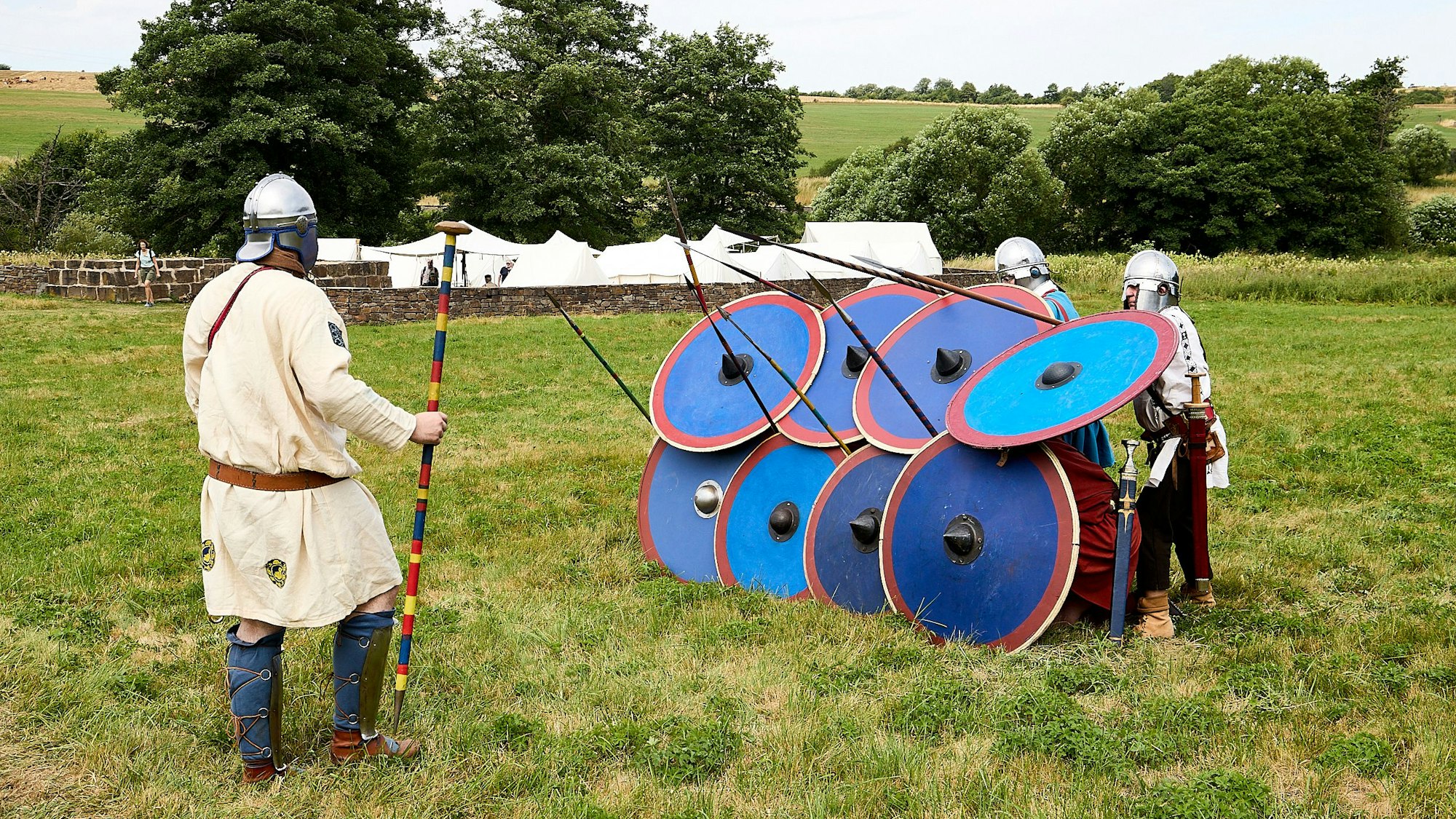 Ein Mann mit Helm steht vor Männern, die ihm hinter runden Schilden Speere entgegenstrecken.