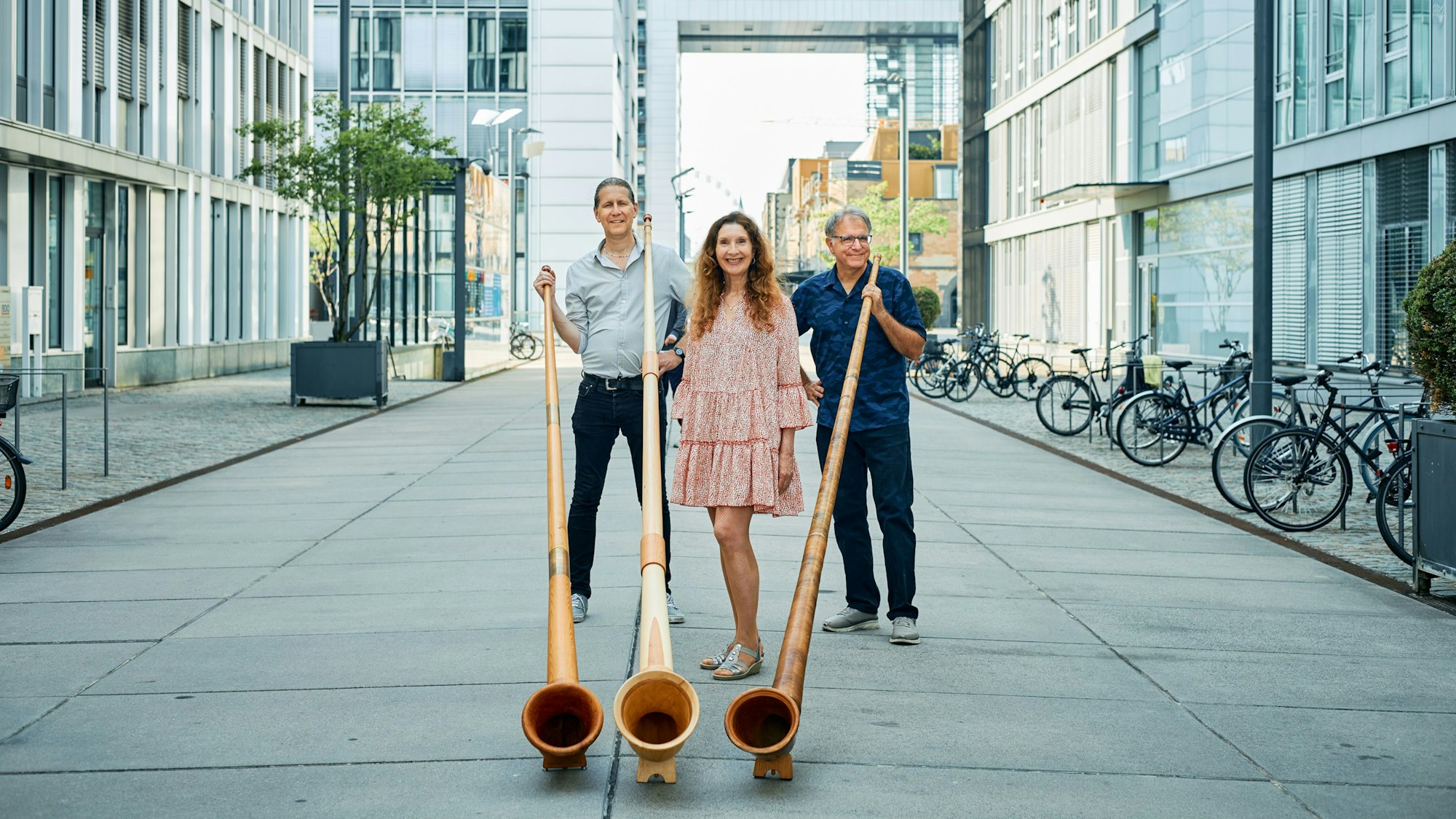 Zwei Frauen und ein Mann stehen mit Alphörnern vor Kranhäusern in einer Stadt.